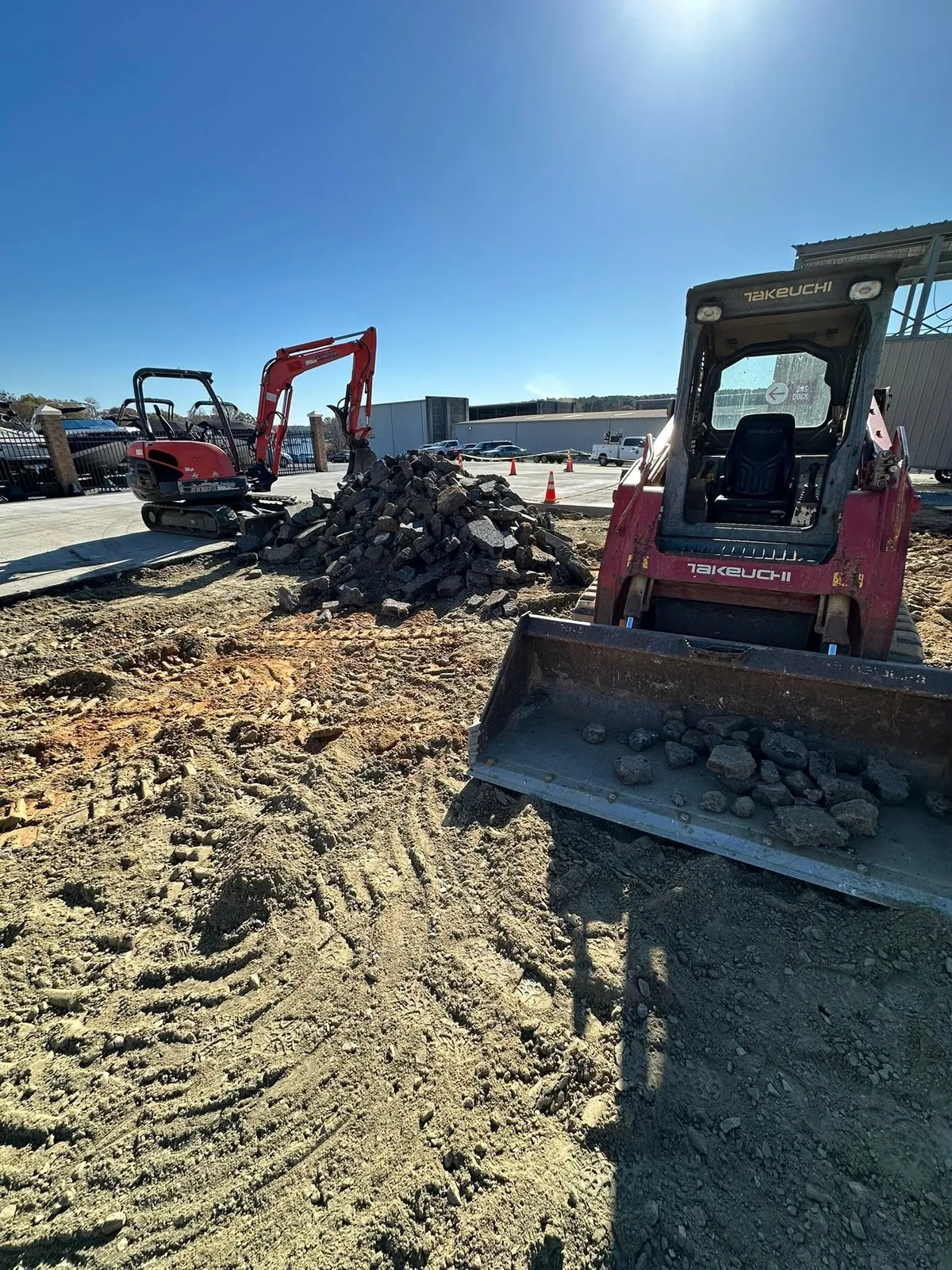 Takeuchi track loader scooping broken asphalt pile, Kubota mini excavator behind, marina lot