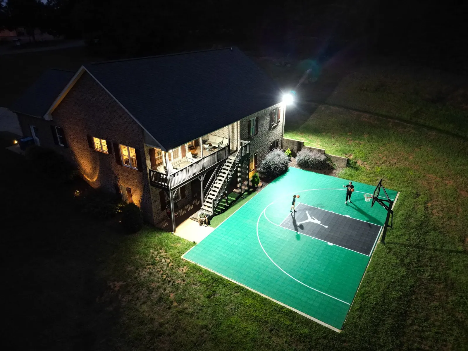 Night drone shot of floodlit backyard basketball court with green tile surface beside two-story brick house