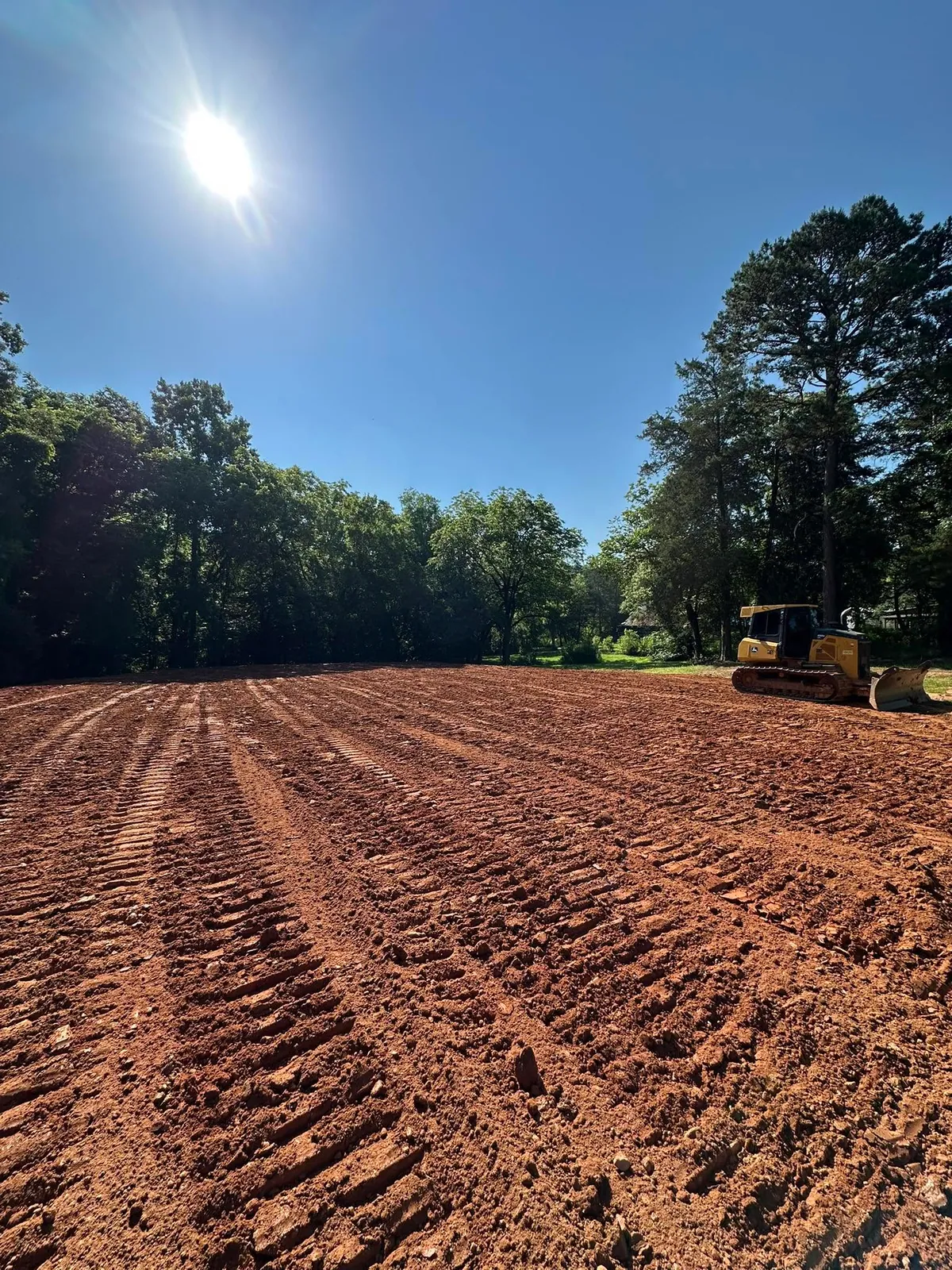John Deere crawler dozer on freshly graded red clay barn pad, bright sun, tree line behind