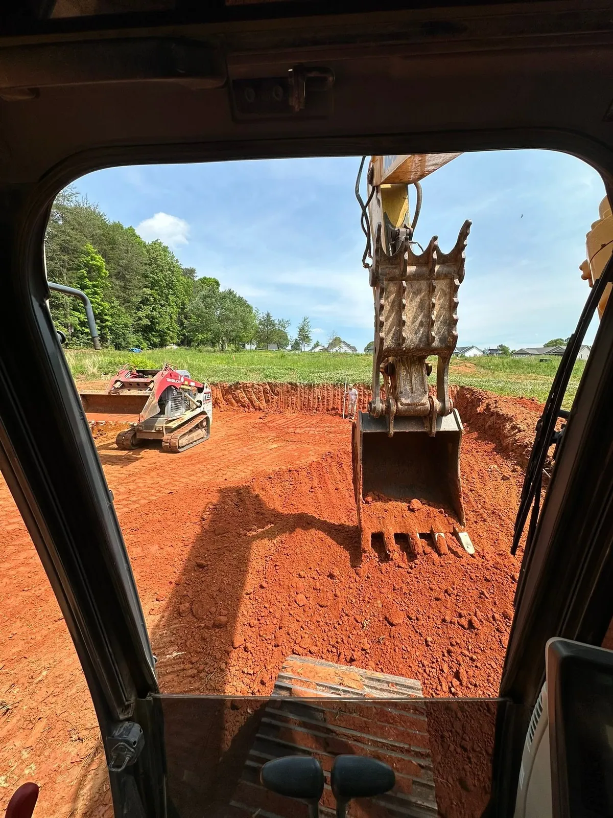 Excavator cab view of bucket cutting red clay pad while Takeuchi track loader spreads spoil, subdivision behind