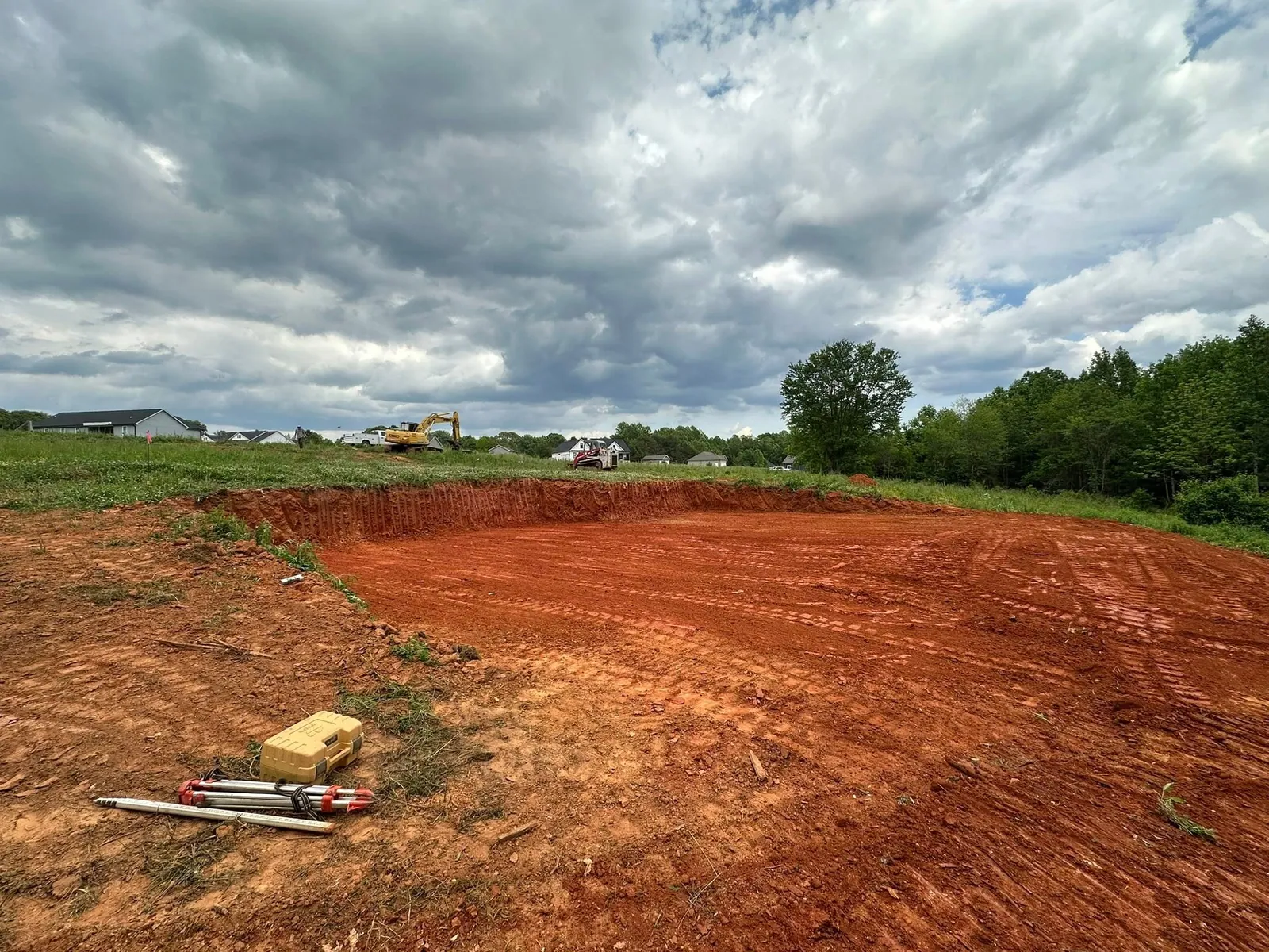 Wide view of finished red clay building pad cut with laser tripod, Kobelco and loader beyond