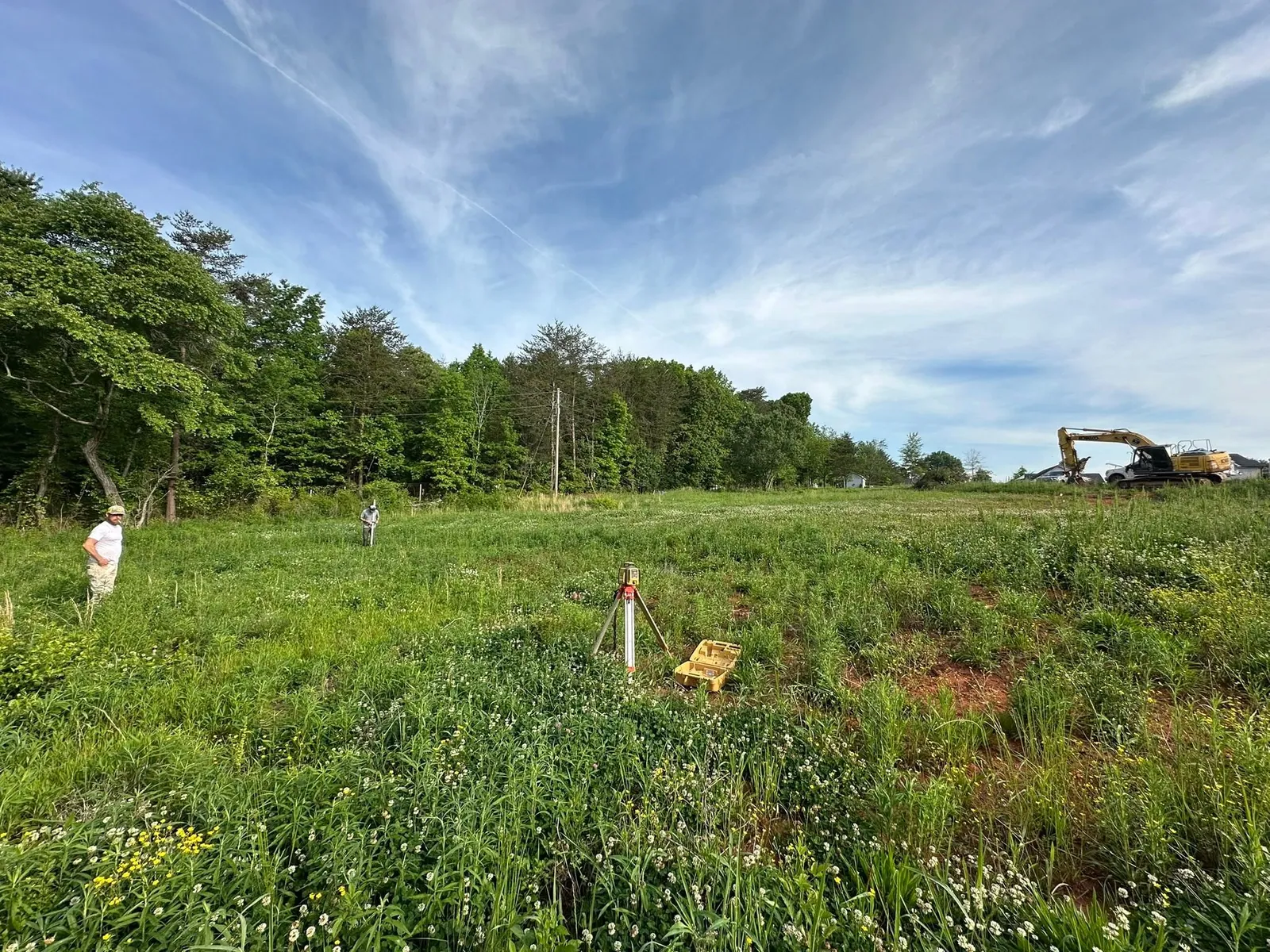 Overgrown field before pad cut, laser level on tripod, crew staking, Kobelco excavator arriving on trailer