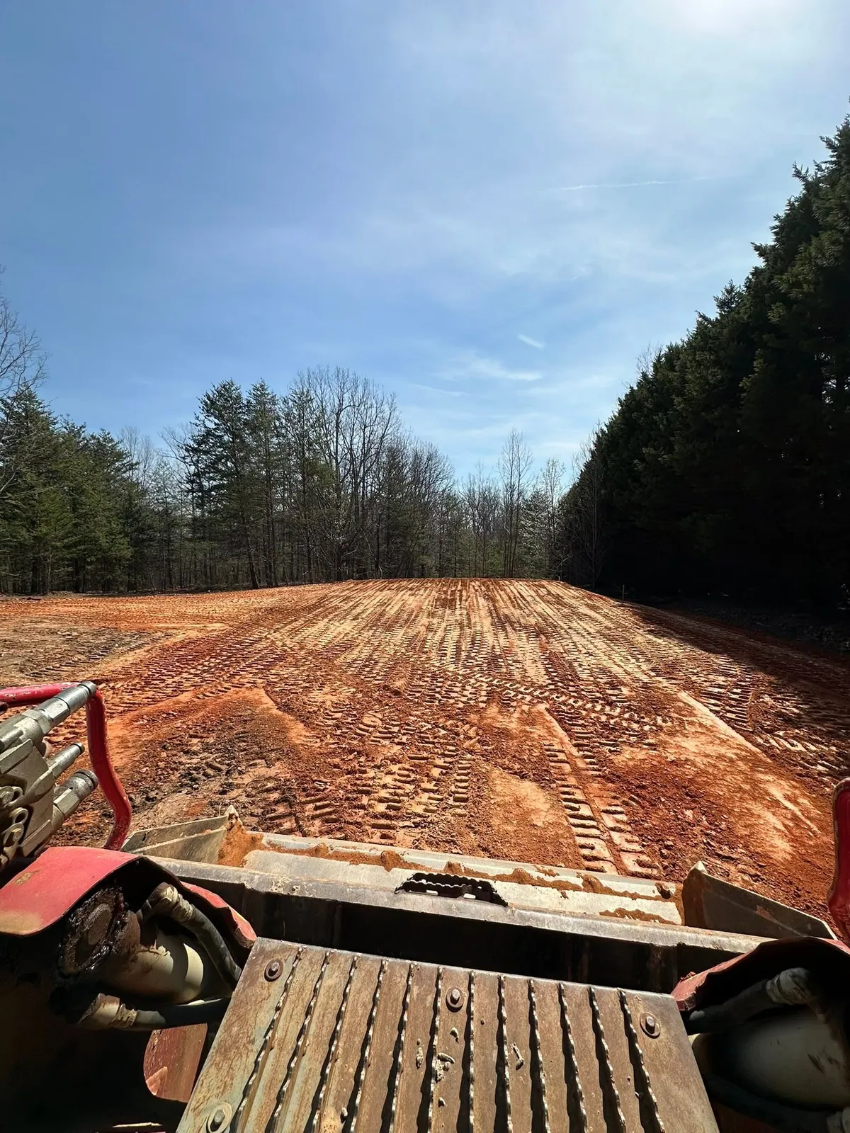 Operator view from track loader down long graded red-clay pad, cypress trees on right