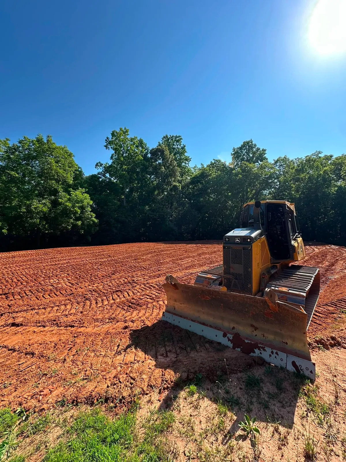 John Deere crawler dozer parked on freshly graded red-clay pad, summer hardwoods behind