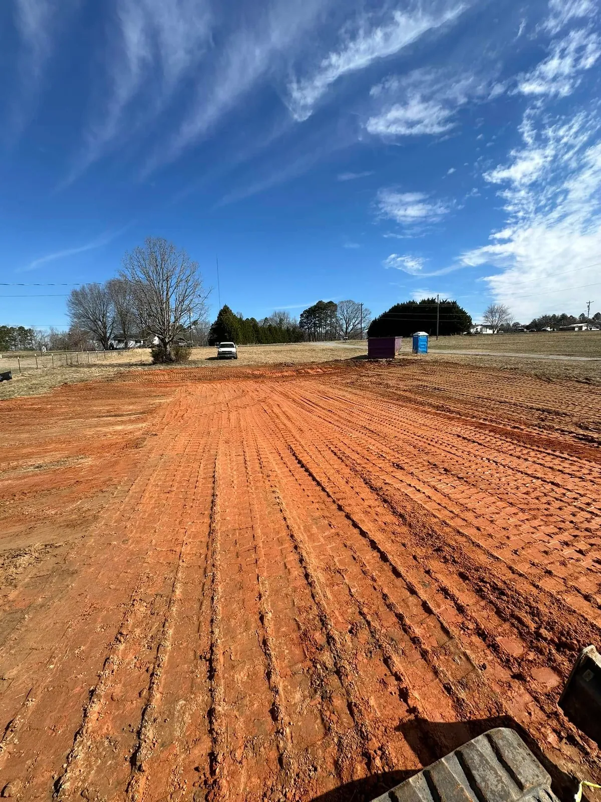 Large red clay building pad track-packed level in open field; roll-off dumpster and porta-john at edge
