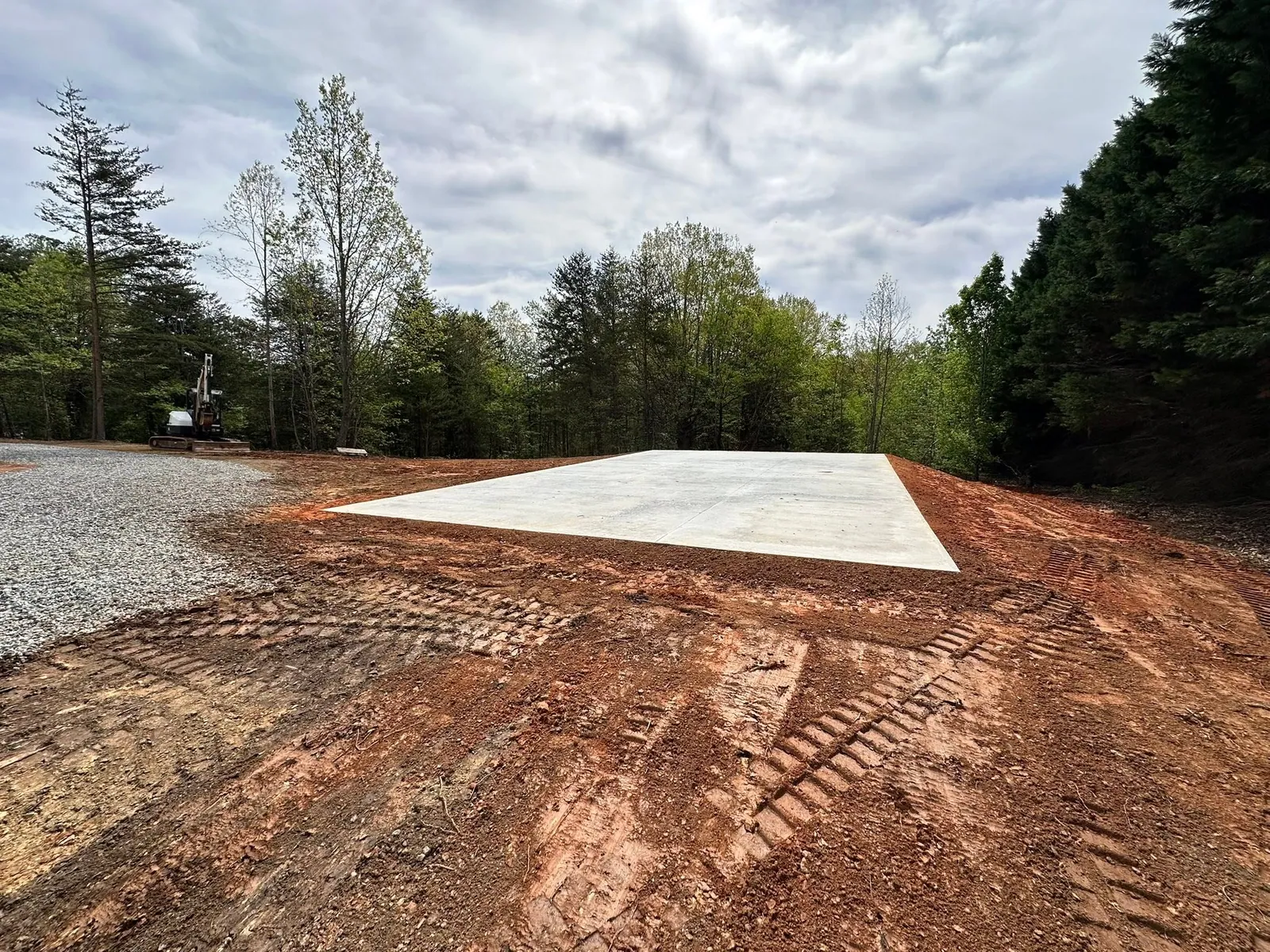 Cured concrete slab on graded red-clay pad in wooded lot, gravel drive at left