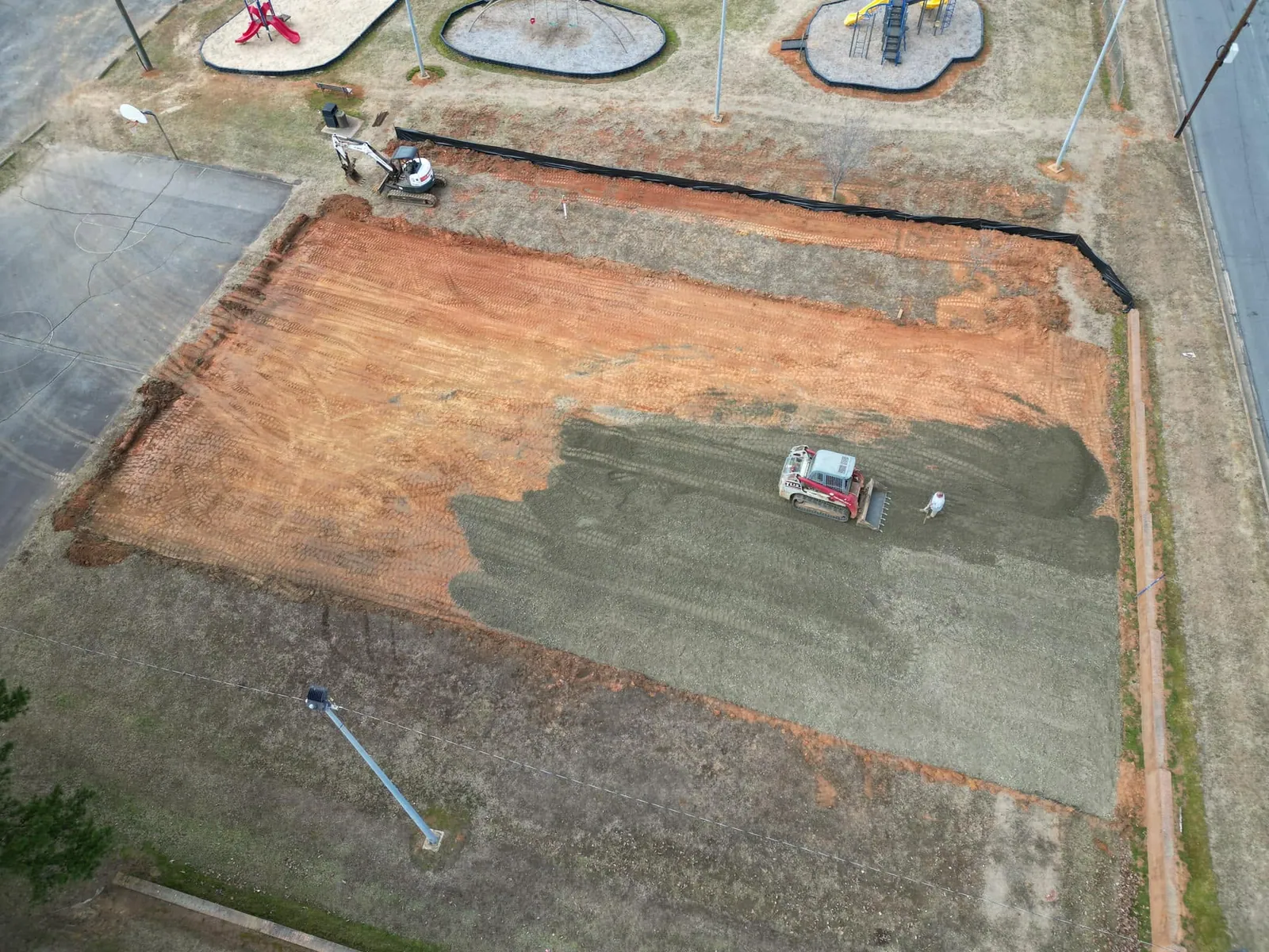 Drone view of rectangular graded pad, gravel base half spread; Takeuchi track loader and mini excavator beside playground in Maiden, NC