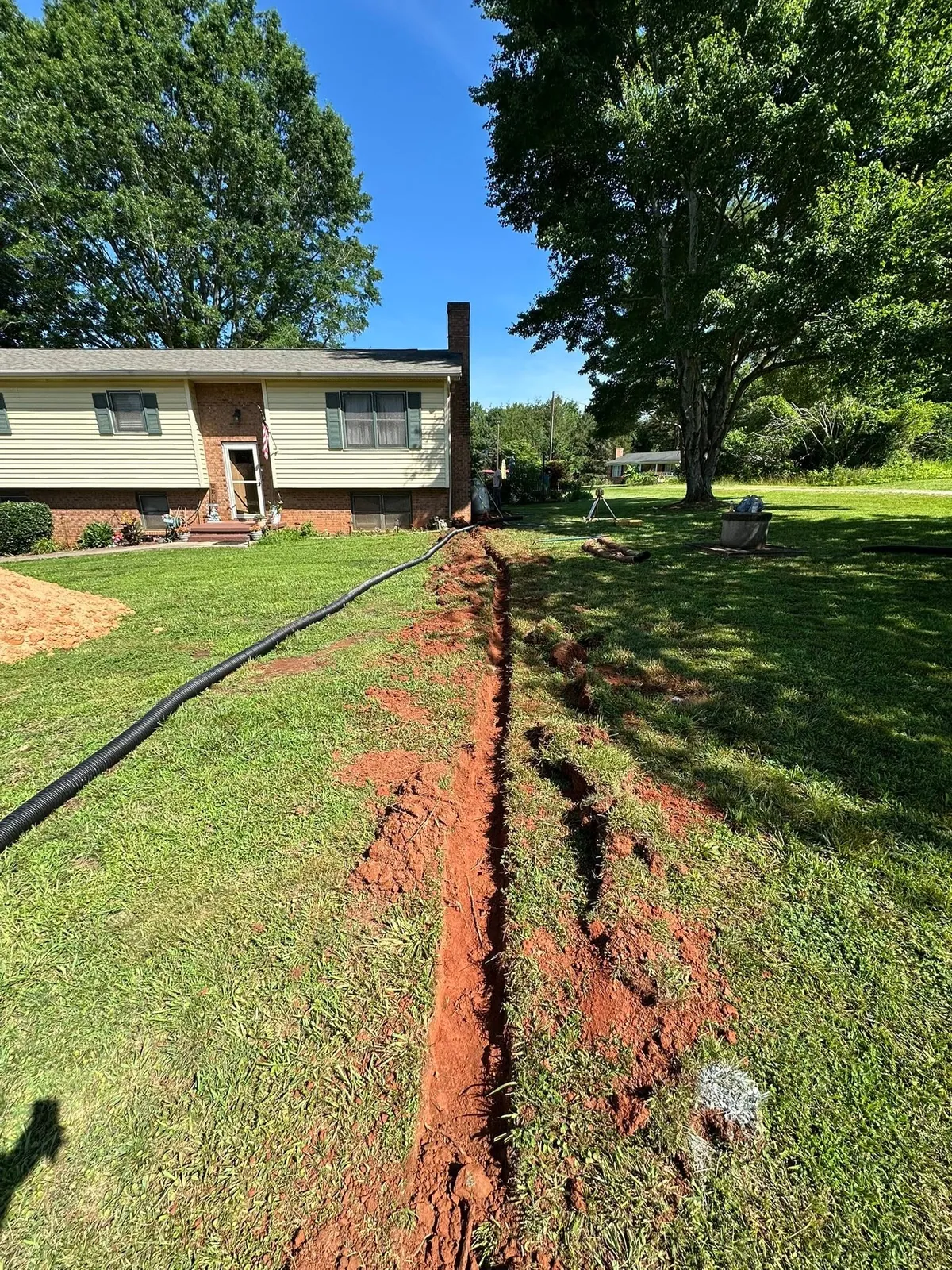 Trench cut across front lawn with black corrugated drain pipe laid, split-level house behind