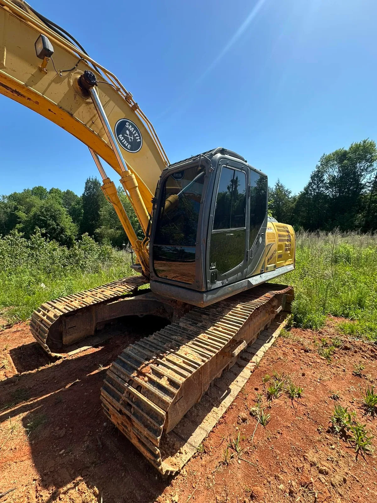 Low-angle glamour shot of Kobelco excavator with Smith Mine logo on boom, parked on red clay by tall grass
