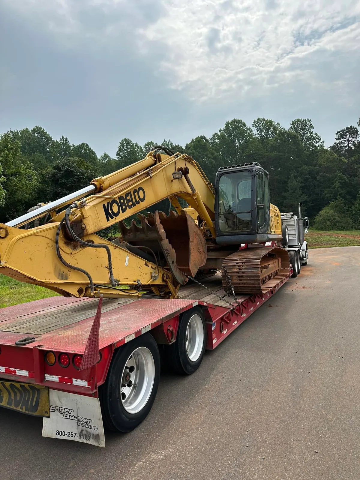 Yellow Kobelco excavator with hydraulic thumb chained on red Eager Beaver lowboy behind road tractor