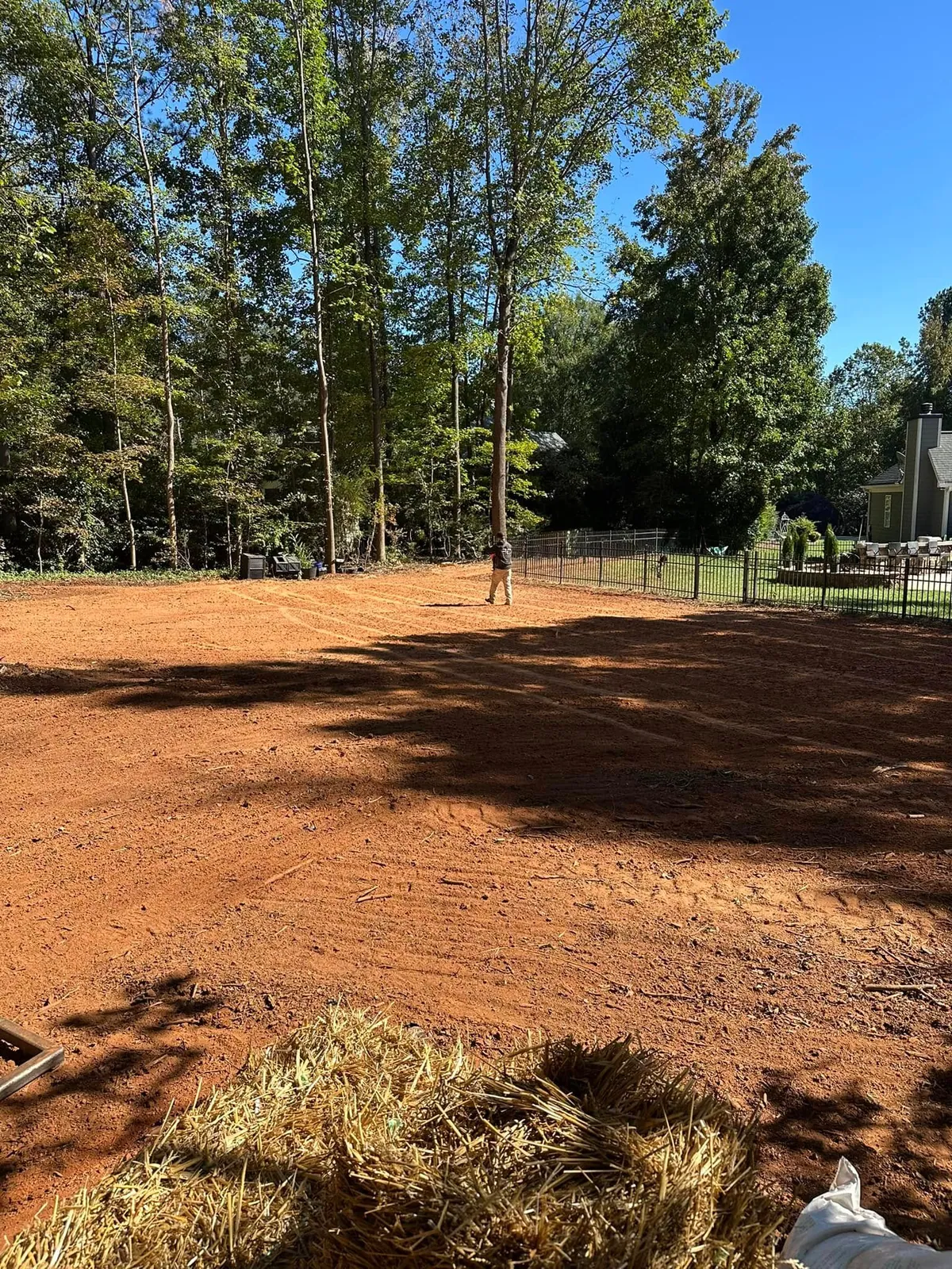 Graded red-clay backyard with straw bales staged for seeding, worker walking, black fence, patio