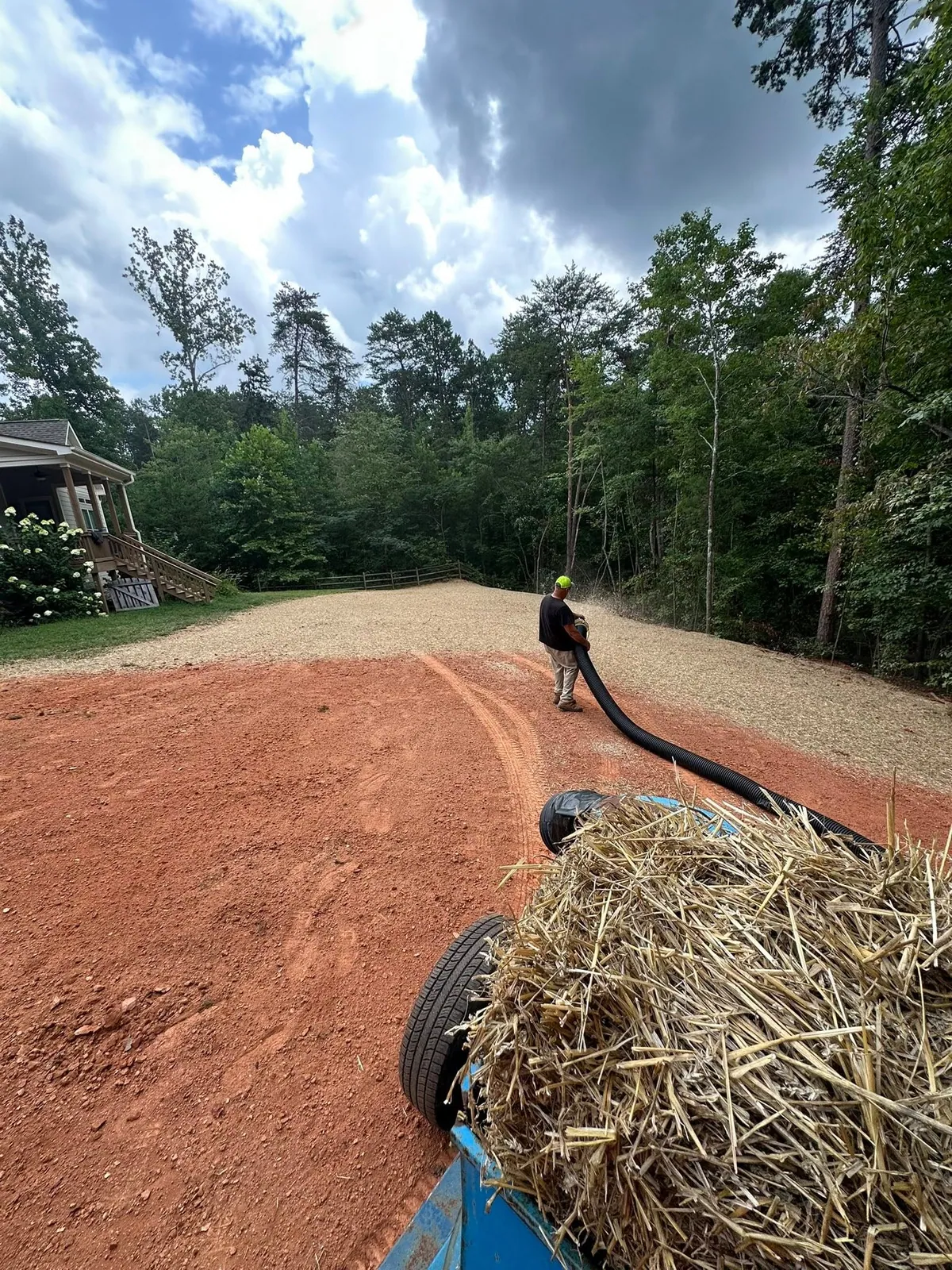 Worker blowing straw over graded backyard fill from trailer-mounted straw blower behind cream house