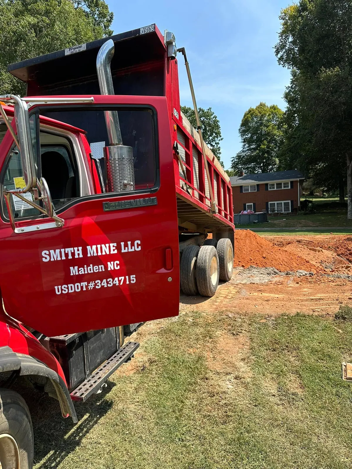 Red Ford Louisville dump truck with bed raised, dumping red clay fill in yard of brick ranch house