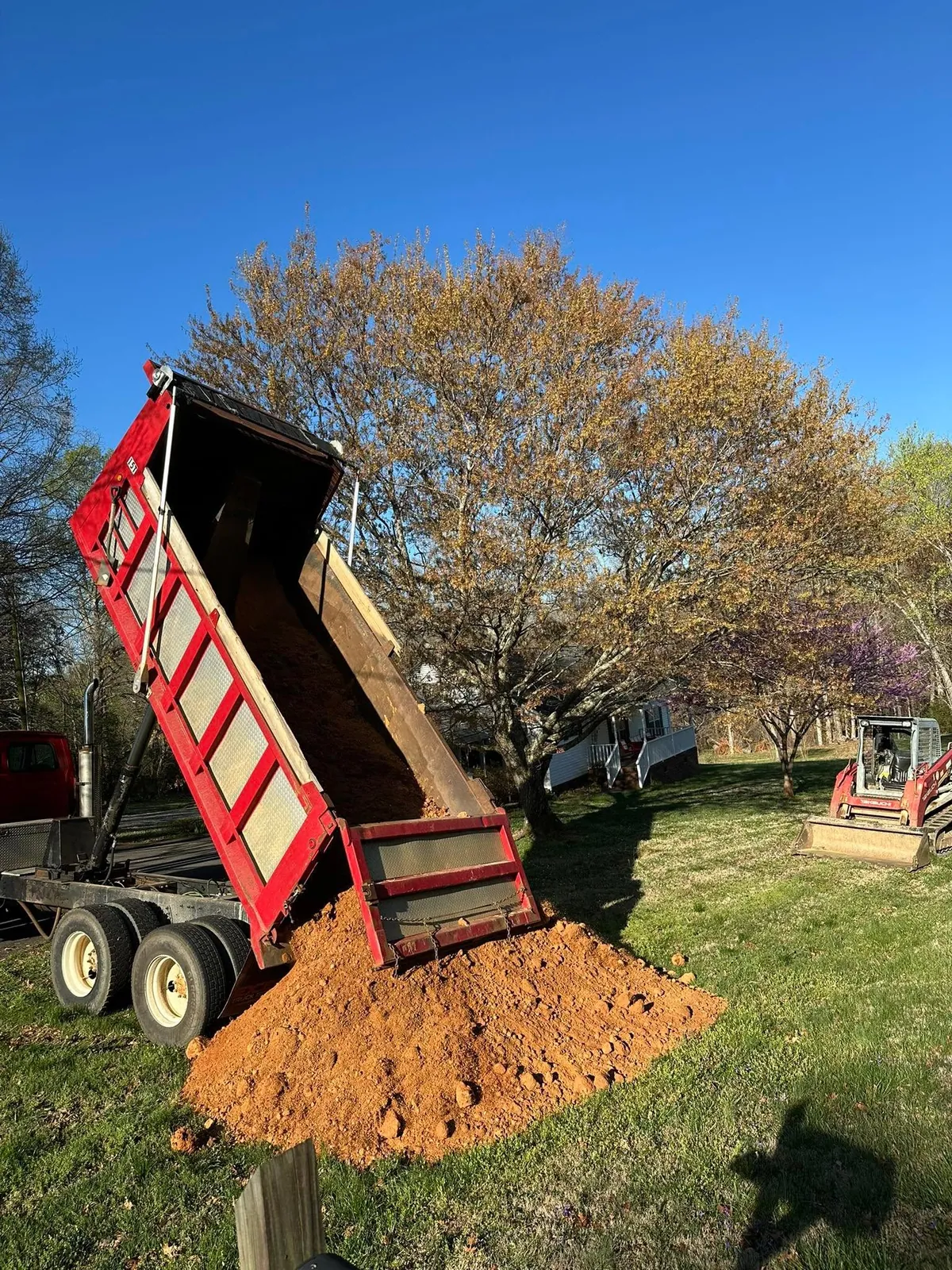 Red dump truck bed fully raised dumping red clay fill on lawn; Takeuchi track loader waiting behind