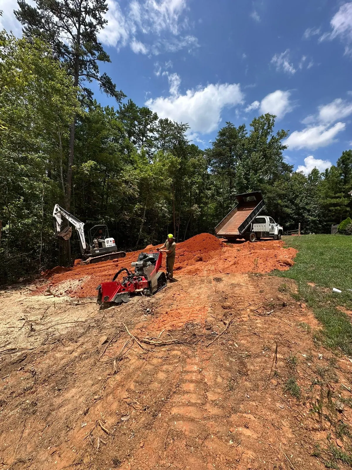 Dump truck tipping red clay fill while Bobcat E50 excavator spreads; worker running stand-on stump grinder