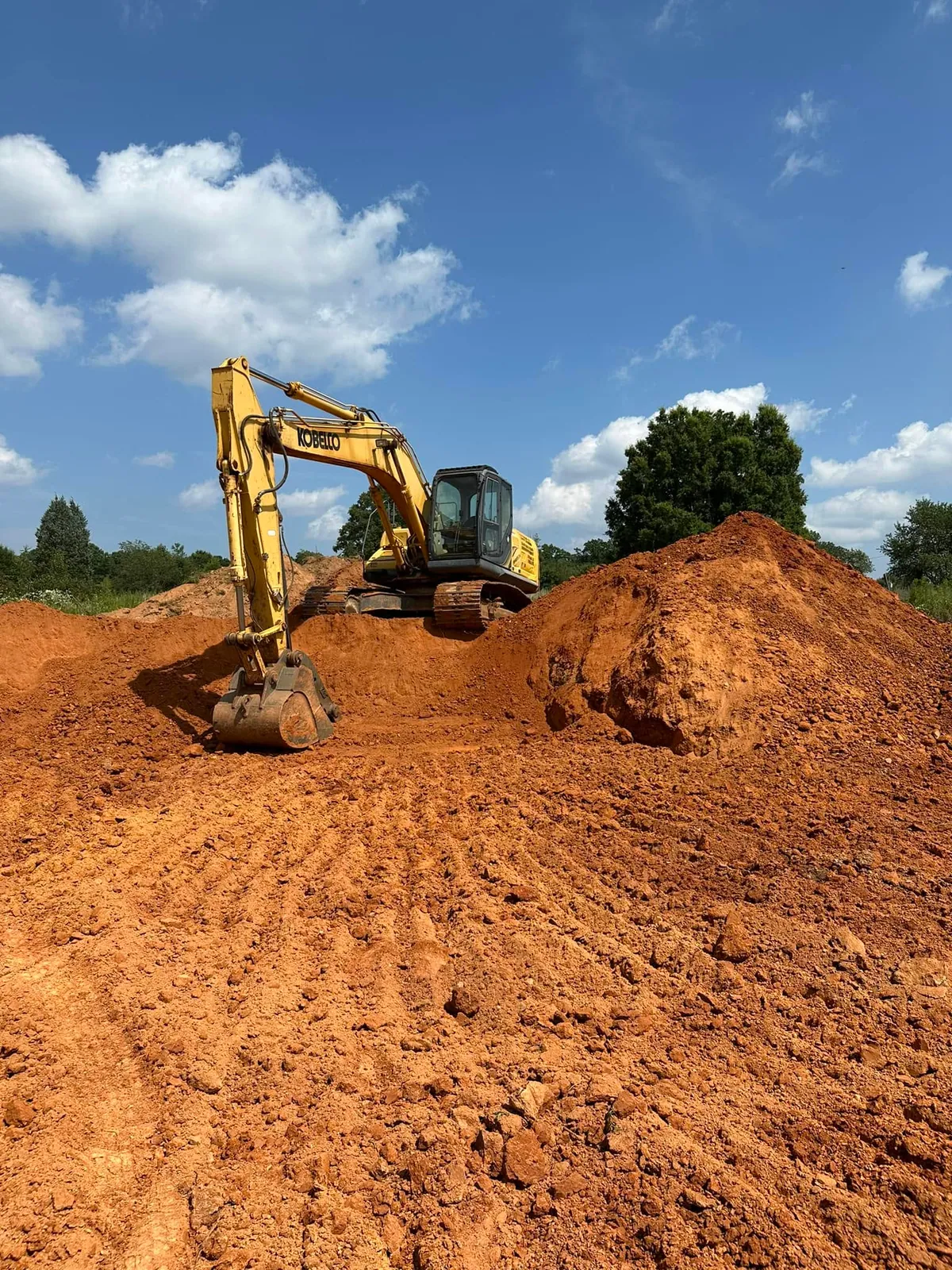 Yellow Kobelco excavator perched atop a large red-clay stockpile under blue sky, bucket resting on cut slope