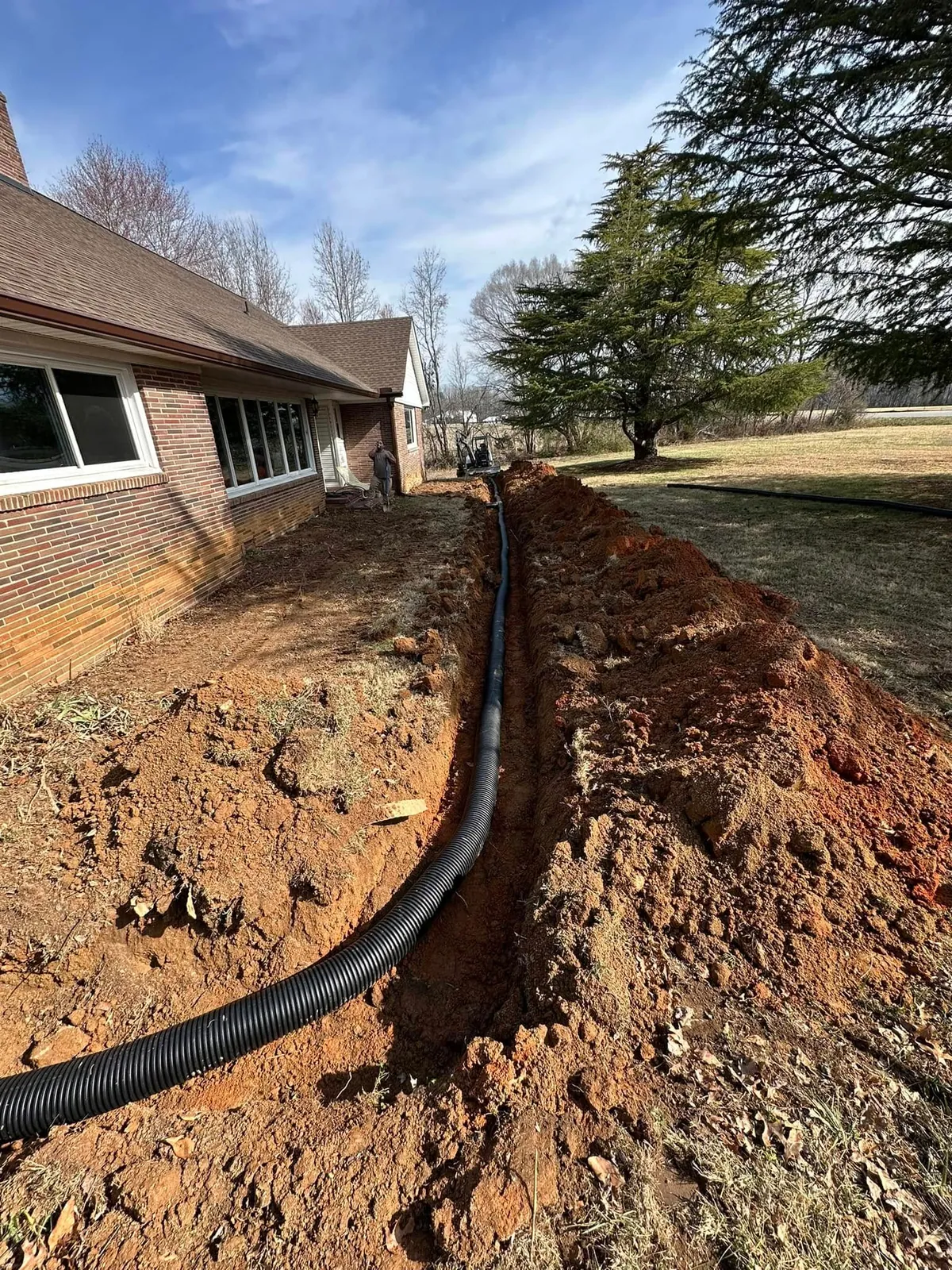Trench dug along brick ranch house with black corrugated drain pipe laid; mini excavator at far end