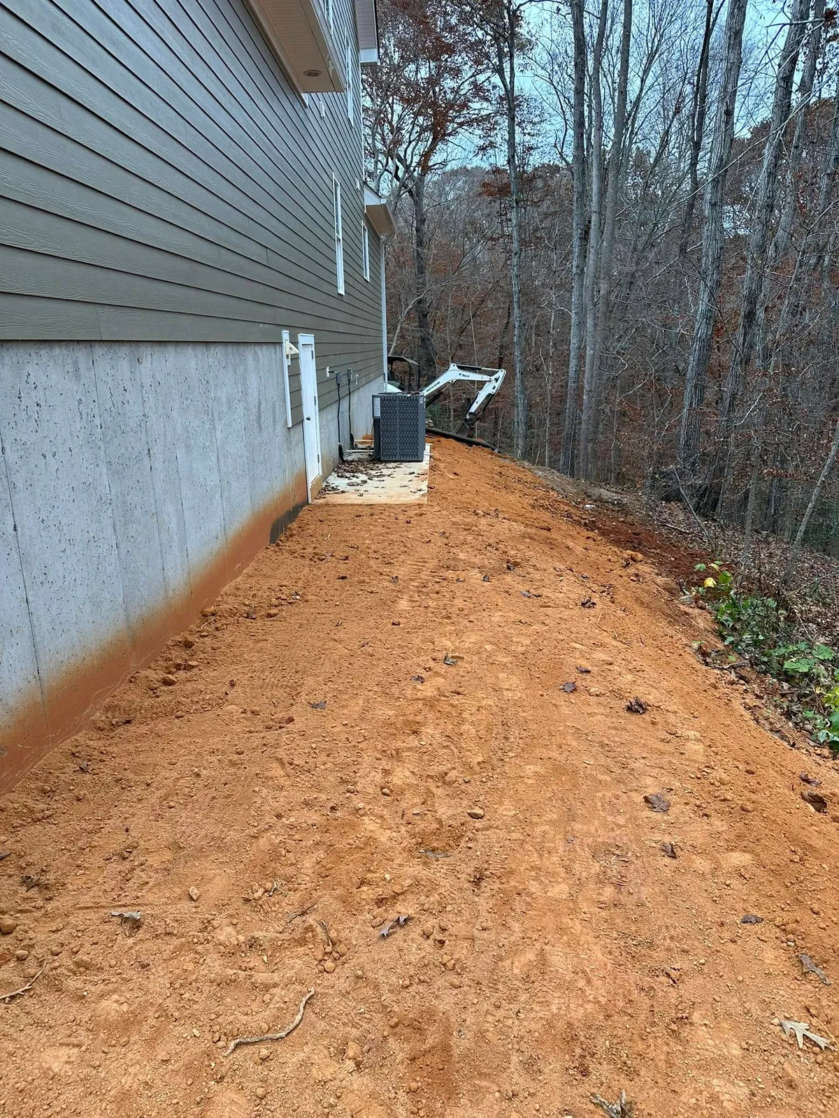 Backfilled and graded strip along new house foundation, white Bobcat mini excavator finishing at far end