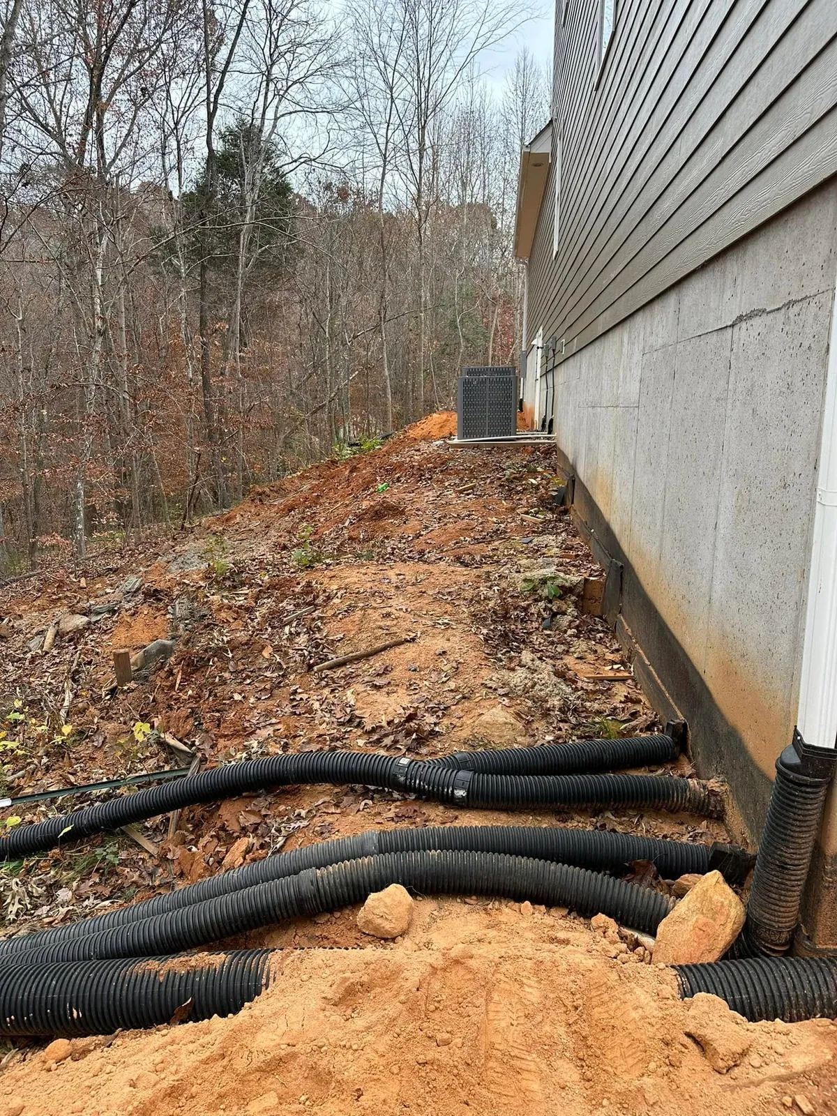Black corrugated drain pipes laid along house foundation wall on wooded slope before backfill, HVAC unit
