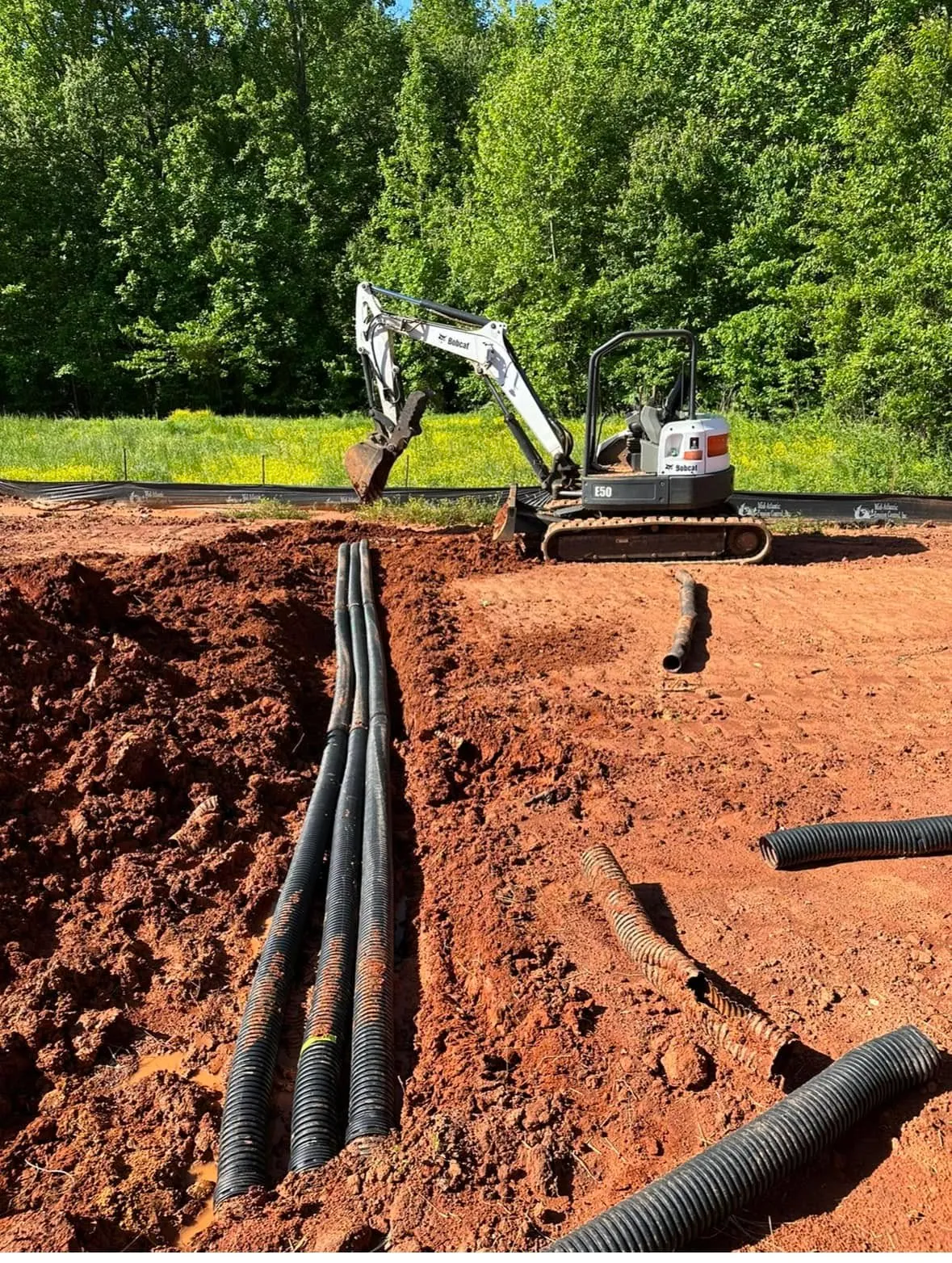 Bobcat E50 mini excavator trenching, twin corrugated drain pipes laid in red clay, silt fence behind