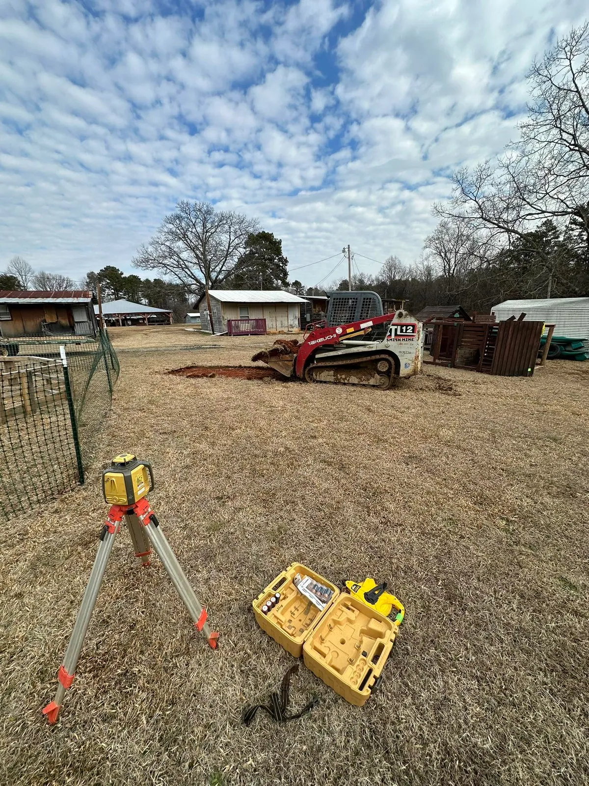 Takeuchi TL12 track loader stripping sod in rural backyard, Topcon laser level on tripod foreground