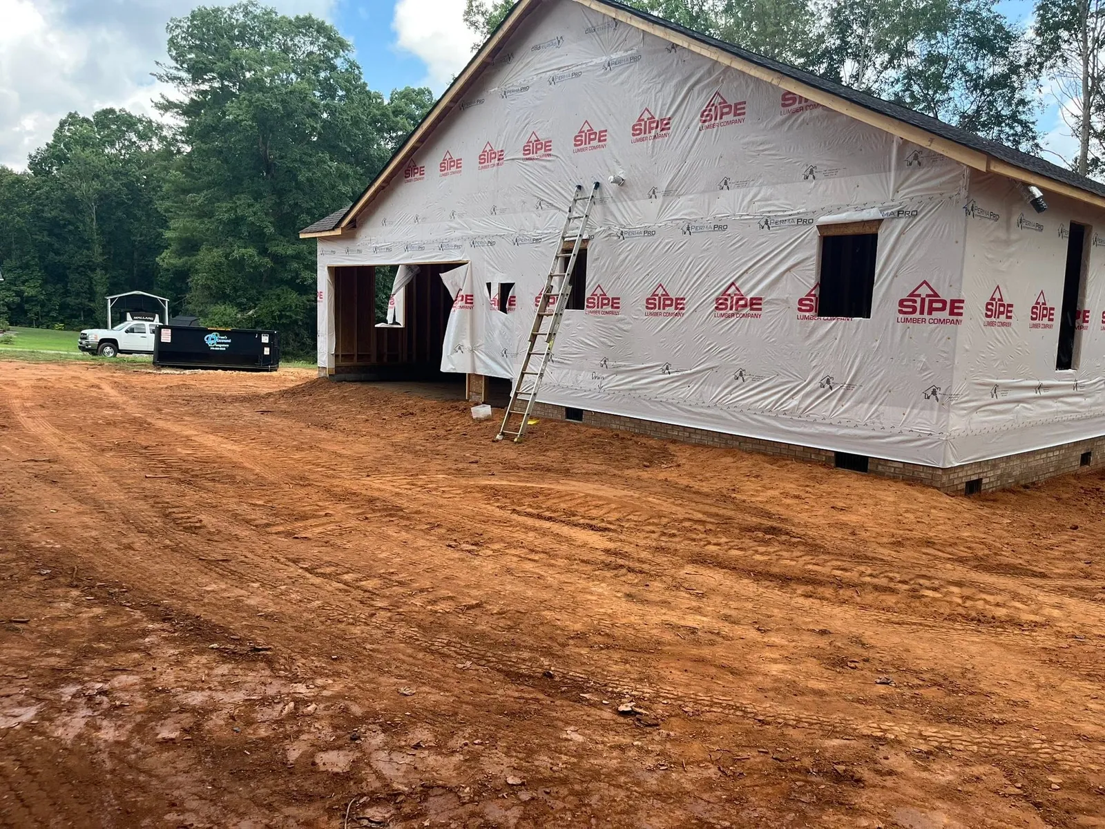 Garage end of housewrapped new build with graded red clay yard, roll-off dumpster and pickup