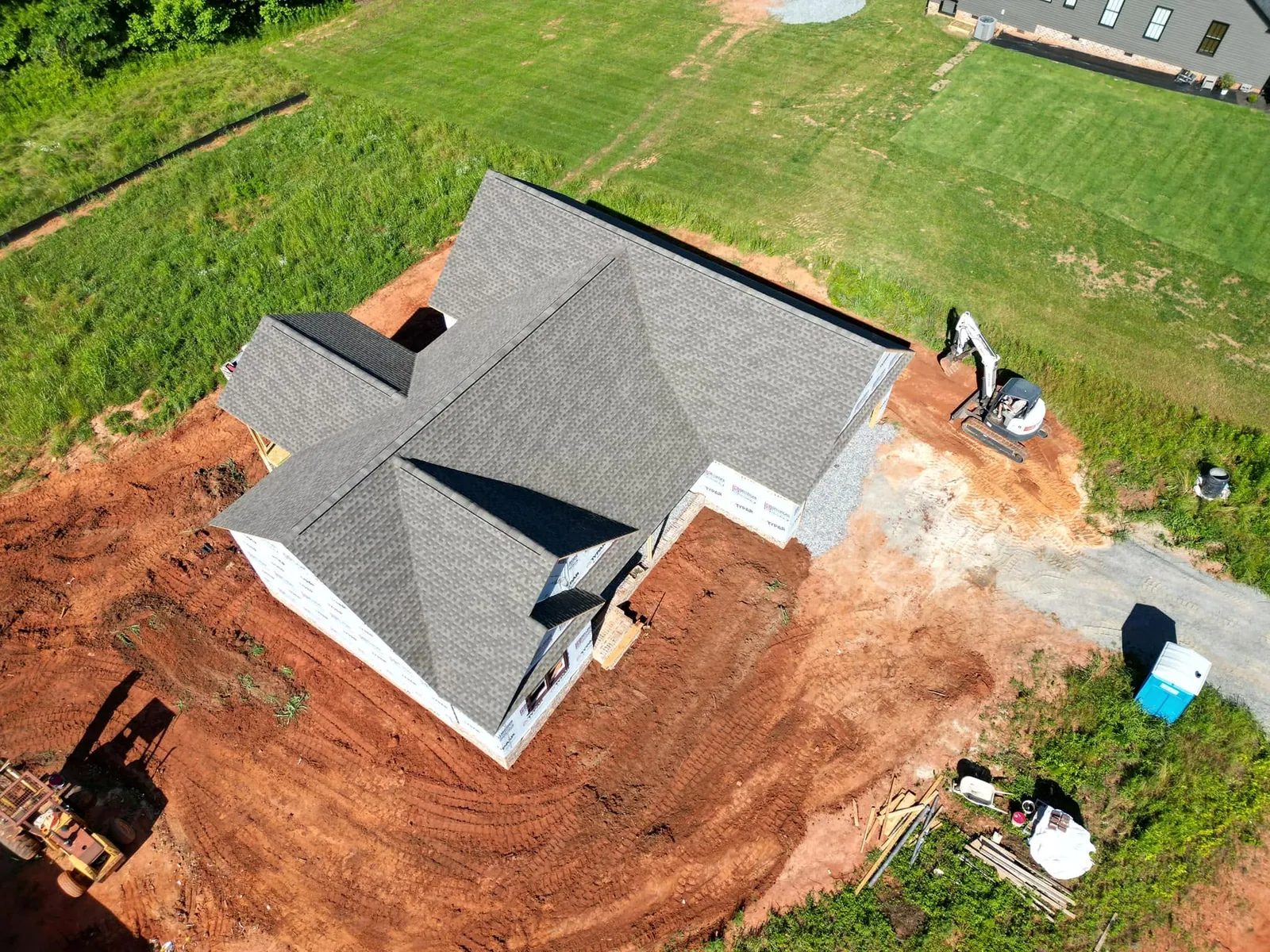 Drone view of newly shingled house with red clay rough-graded around foundation; white mini excavator parked