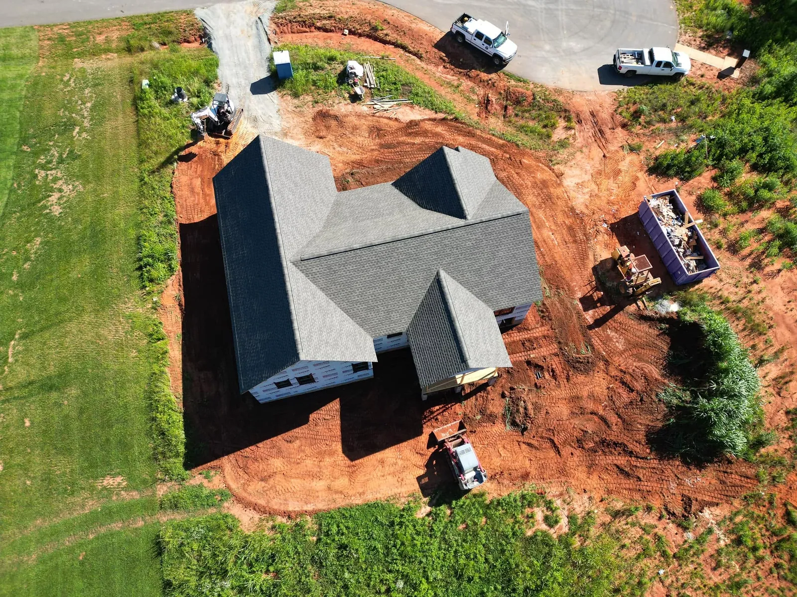 Drone view of new house under construction with red clay rough-graded around it; loader, trucks, dumpster on site