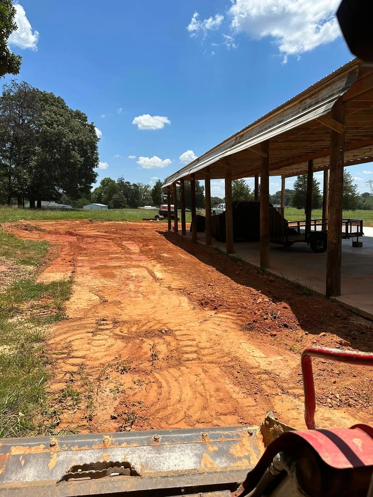 Red clay rough-graded along open pole shed slab, seen from track loader, mini excavator behind