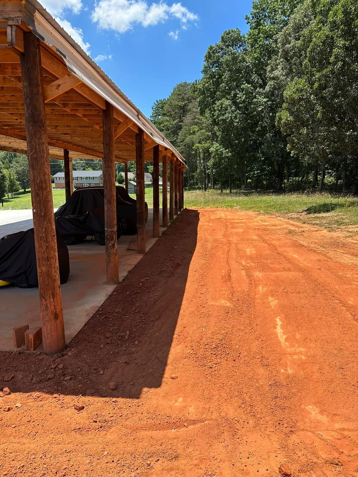 Smoothly graded red clay apron alongside pole shed with covered vehicles, brick house across road