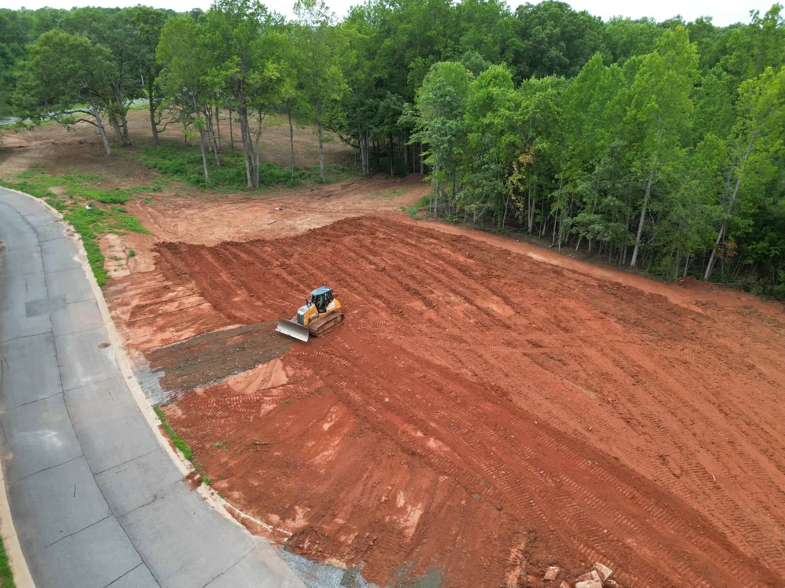 Aerial: dozer grading large red-clay lot beside curbed subdivision road, wooded edge