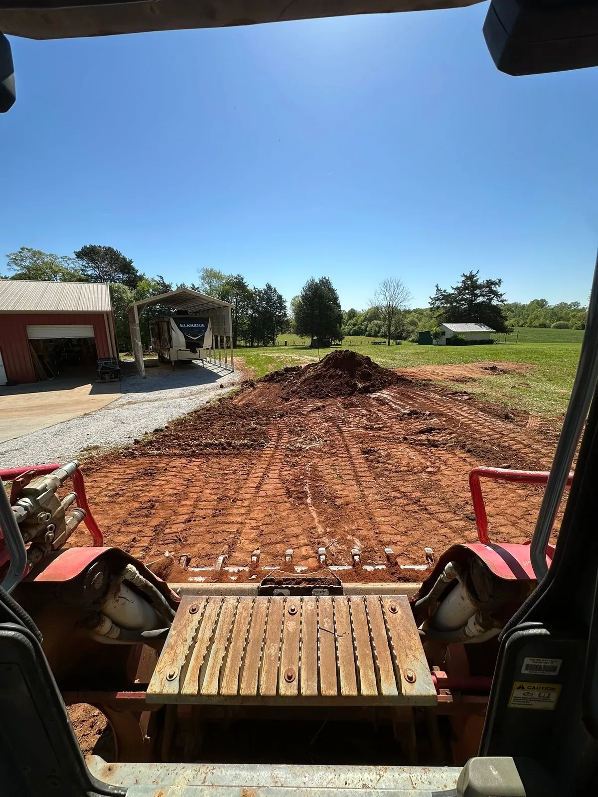 Takeuchi track loader operator view spreading red clay fill pile across yard beside red metal shop