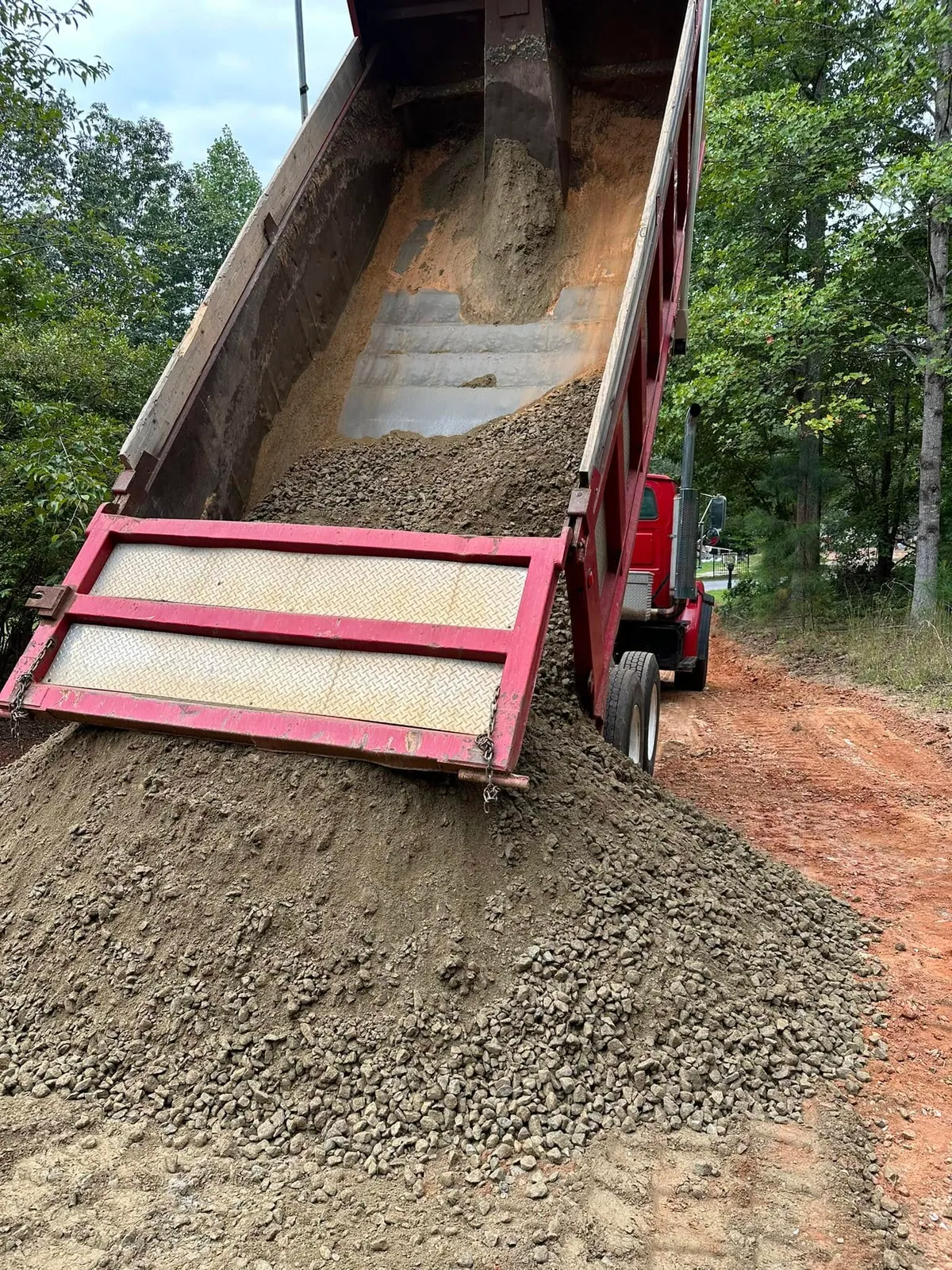 Red tandem dump truck bed raised, tailgate spreading crusher-run gravel down a red-clay wooded driveway