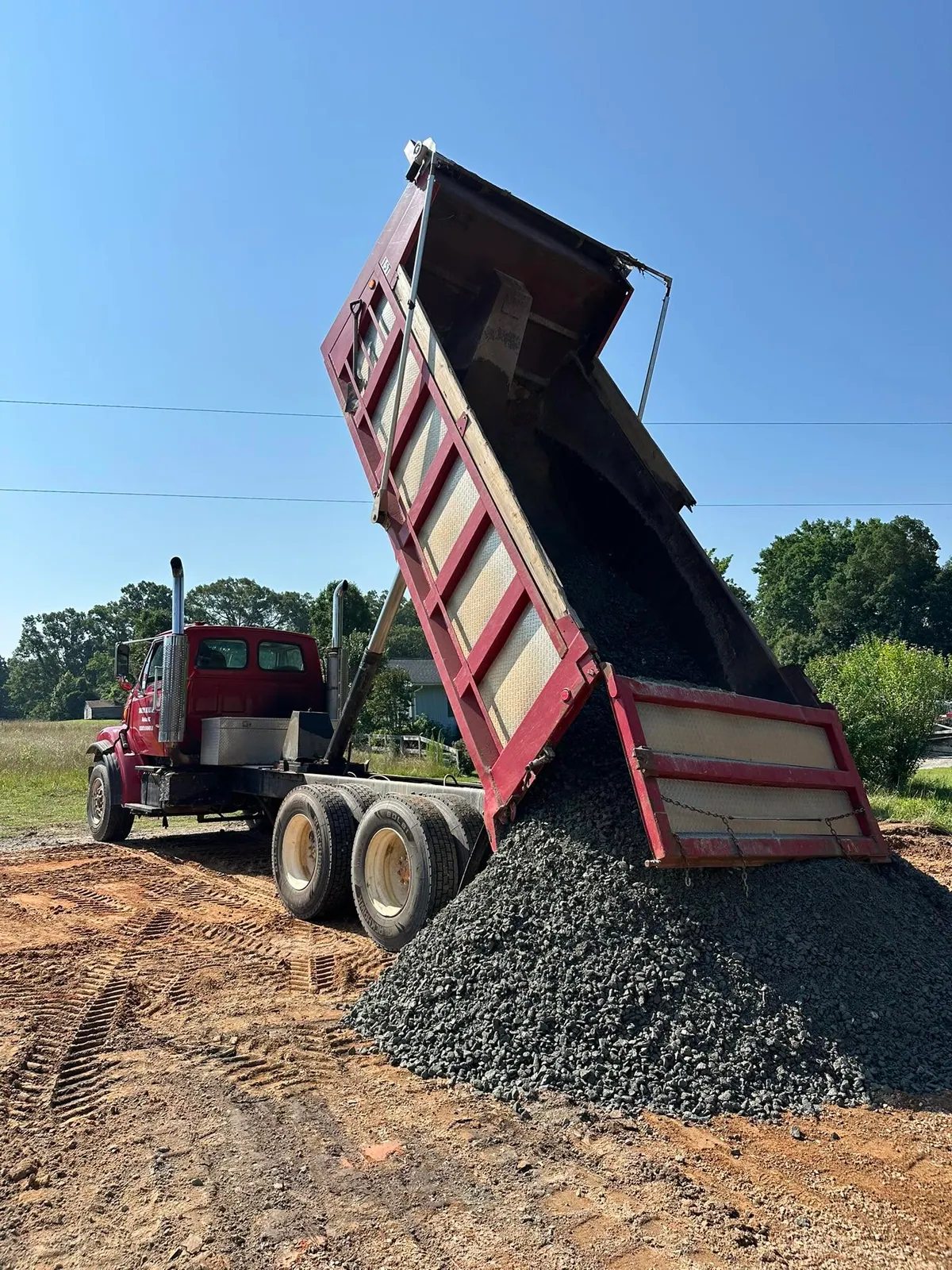 Red tandem dump truck with bed raised dumping gray crushed stone onto graded driveway base