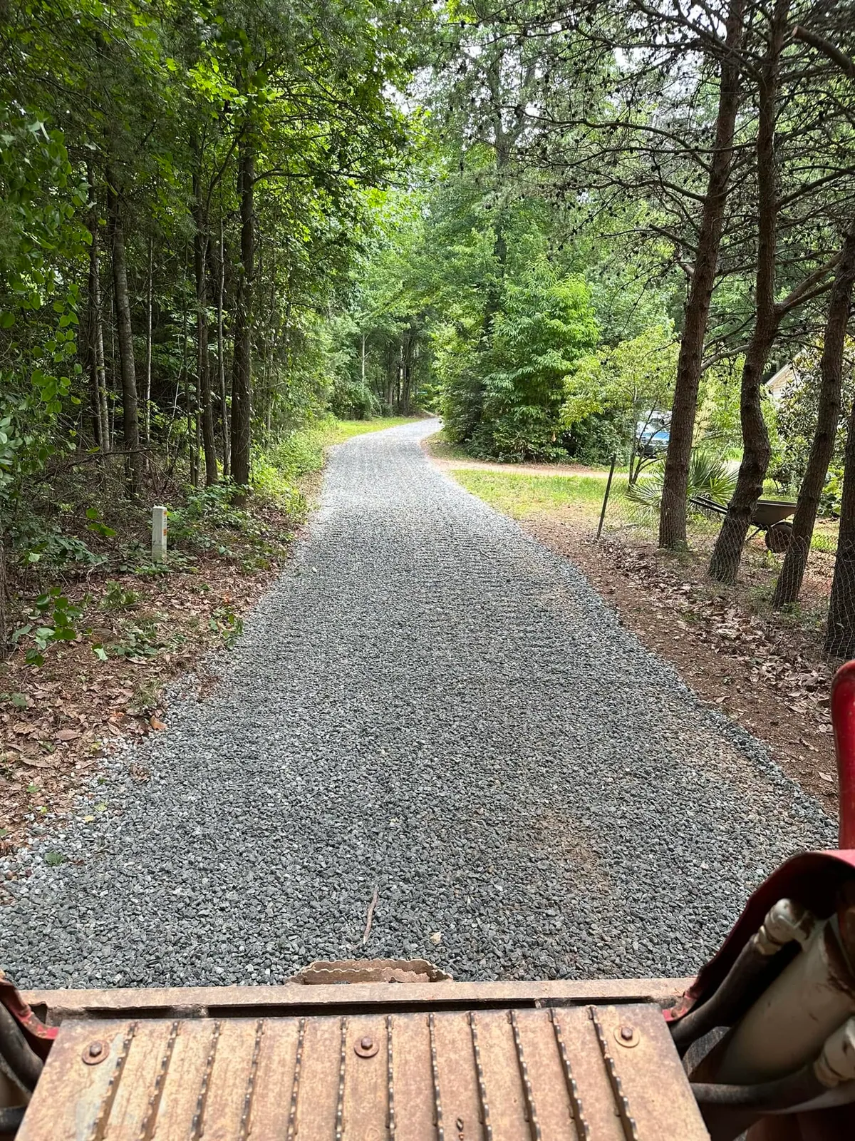 Freshly spread grey crushed stone driveway through woods, seen from red Takeuchi track loader cab