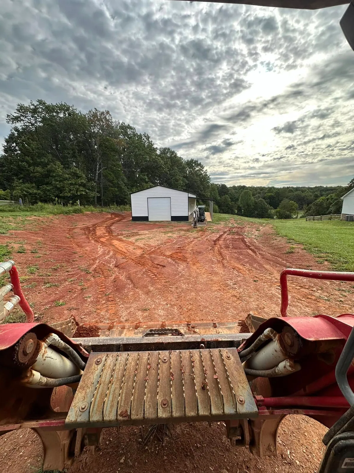 Takeuchi track loader POV over blade: red clay driveway cut toward white metal garage, excavator beyond