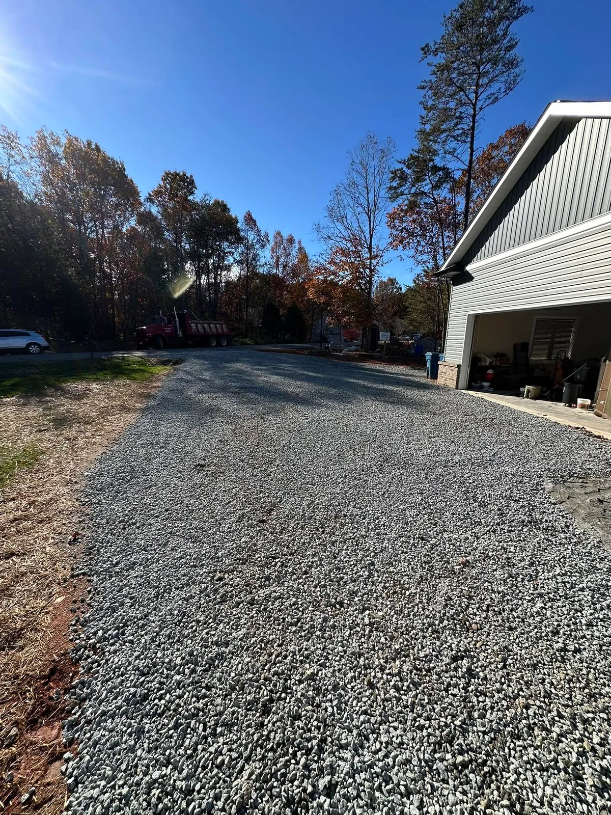 Fresh gray gravel driveway spread beside metal-sided garage, red dump truck parked at road