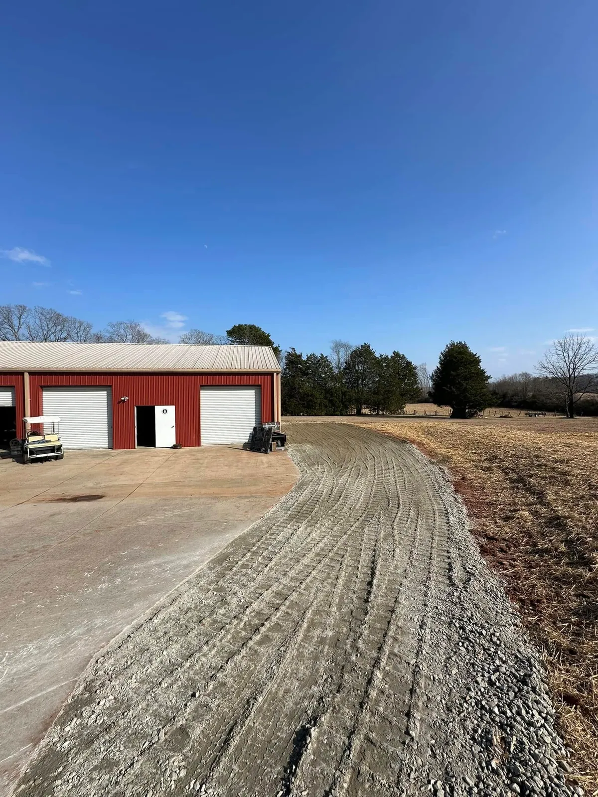 Completed crusher run gravel driveway curving from concrete apron past red metal shop building under clear sky