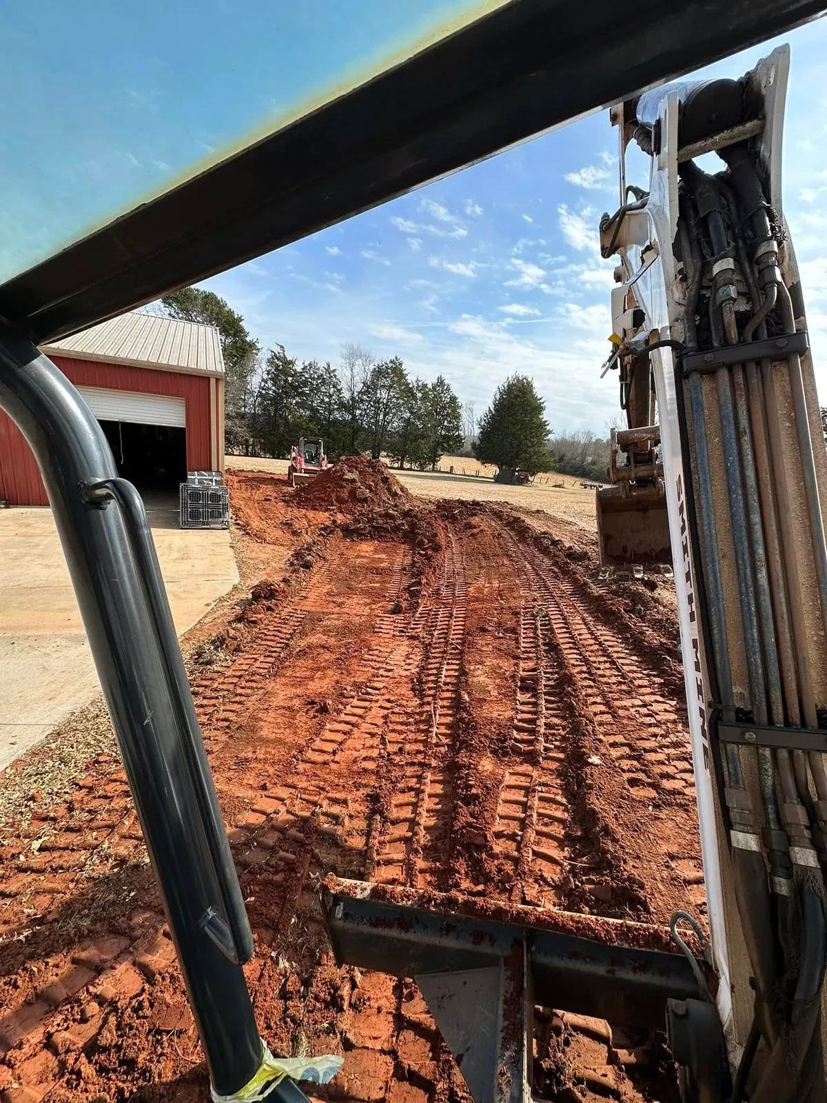 Excavator cab view of red clay driveway being cut beside red barn; Takeuchi track loader pushing spoil pile