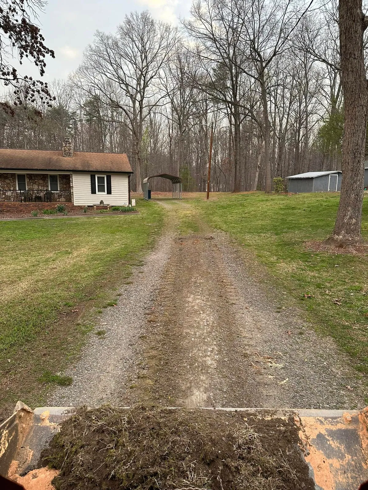 Before shot over loader bucket: thin worn gravel driveway with grass strip toward carport and ranch house
