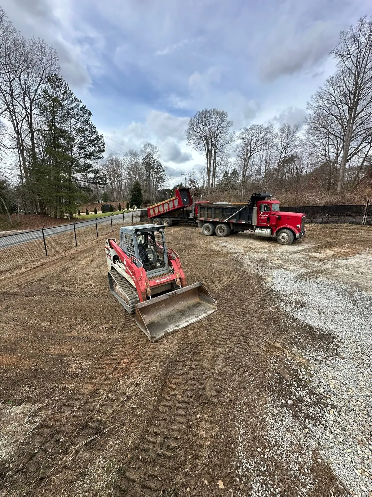 Takeuchi TL12 track loader spreading gravel on graded lot, two red dump trucks dumping behind