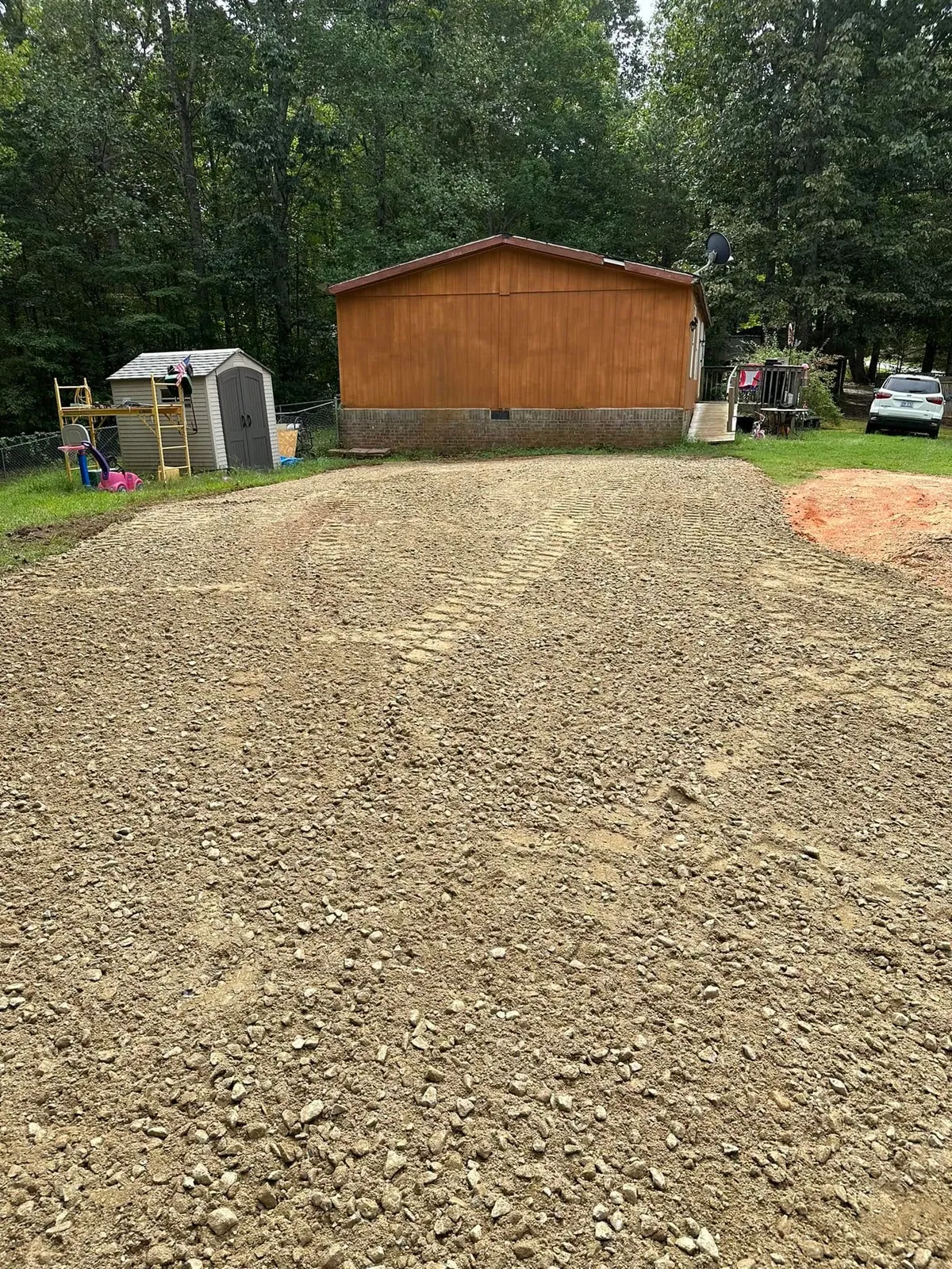 Freshly spread and graded crusher-run gravel parking pad beside wood-sided doublewide and shed