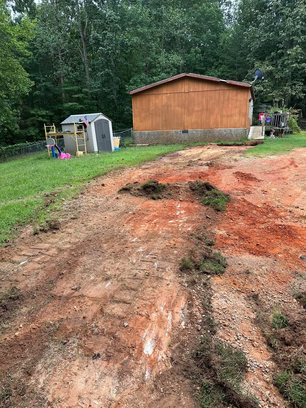 Muddy torn-up red clay yard beside wood-sided doublewide before gravel pad, shed and toys nearby