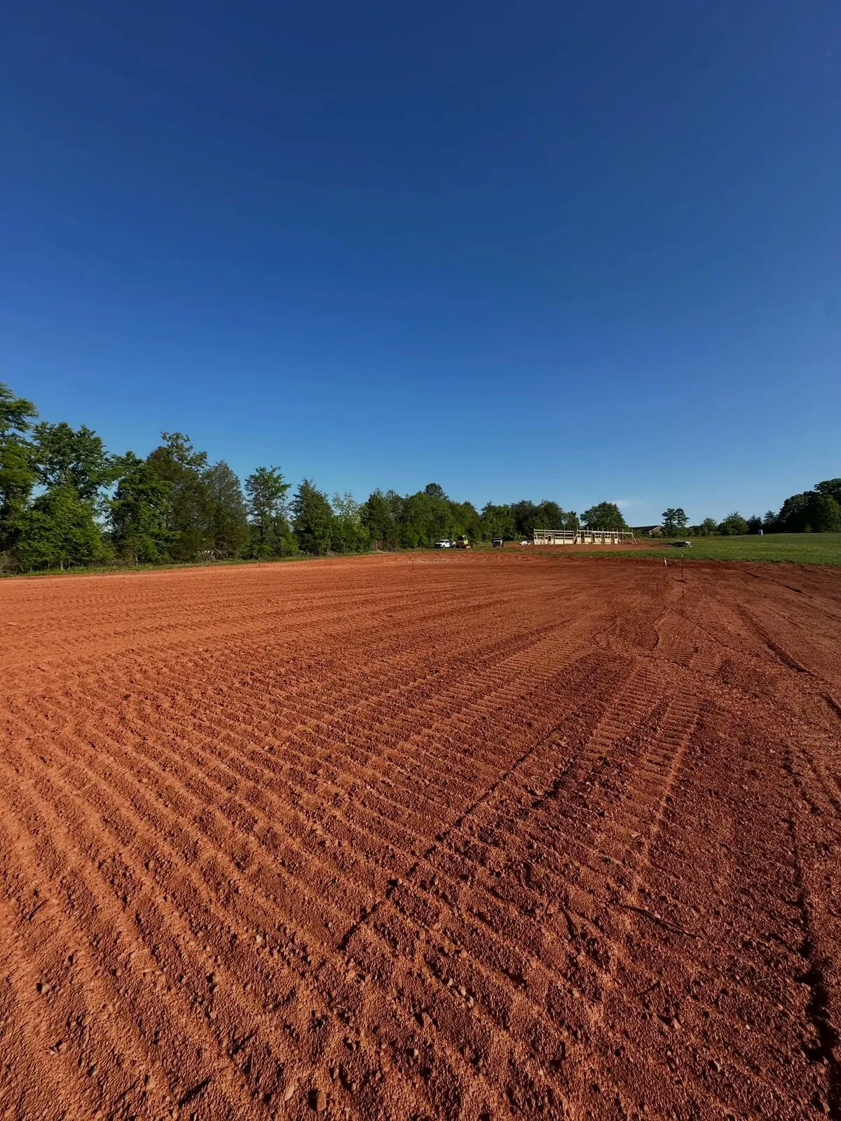 Finished 125x200 horse arena, freshly tracked red clay pad under clear blue sky, barn frame beyond in Claremont, NC