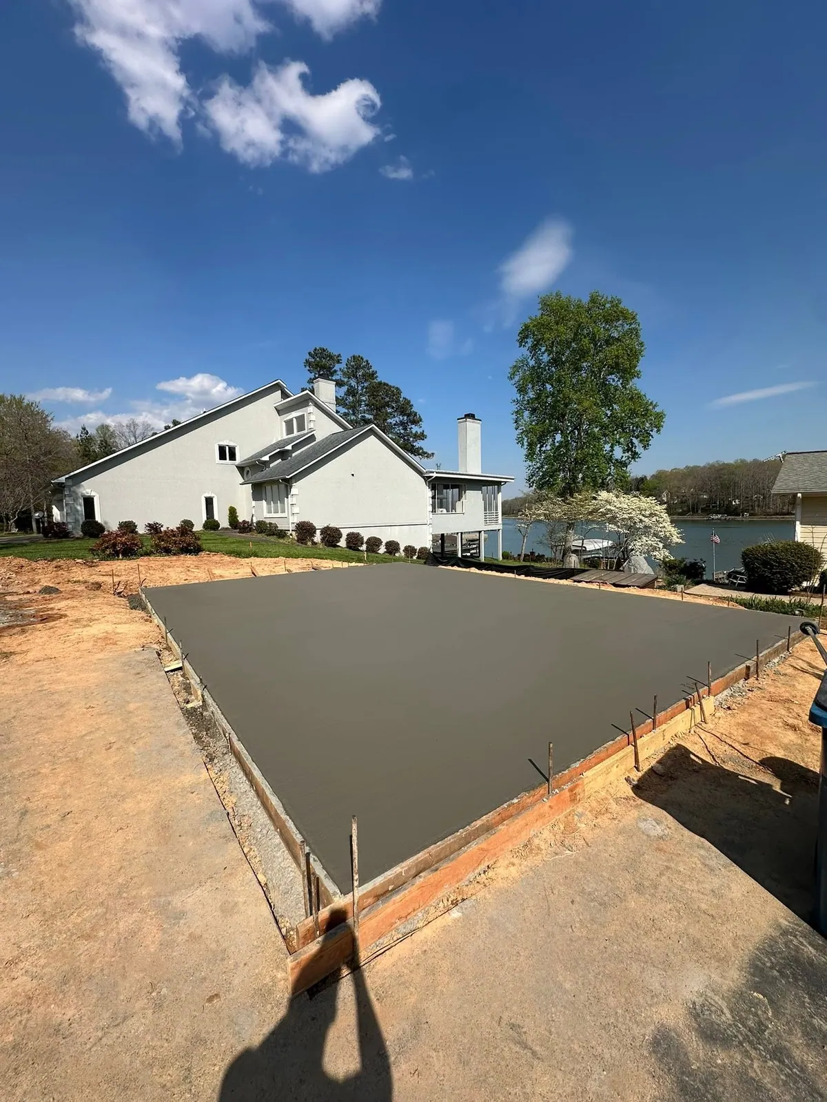 Freshly poured concrete slab in forms beside stucco lakefront house, lake and dogwood behind