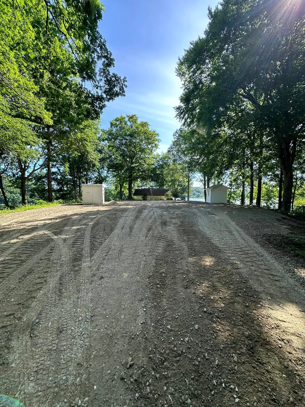 Regraded gravel pad on lakeside lot with fresh track marks; container and shed on either side, lake behind