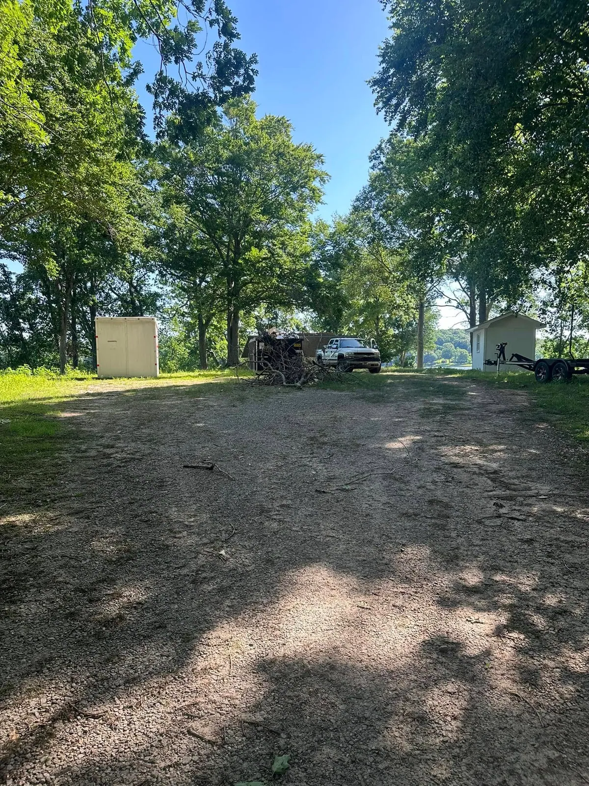 Wooded lakeside lot before work: bare patchy dirt, cargo trailer, brush pile, white pickup, small shed