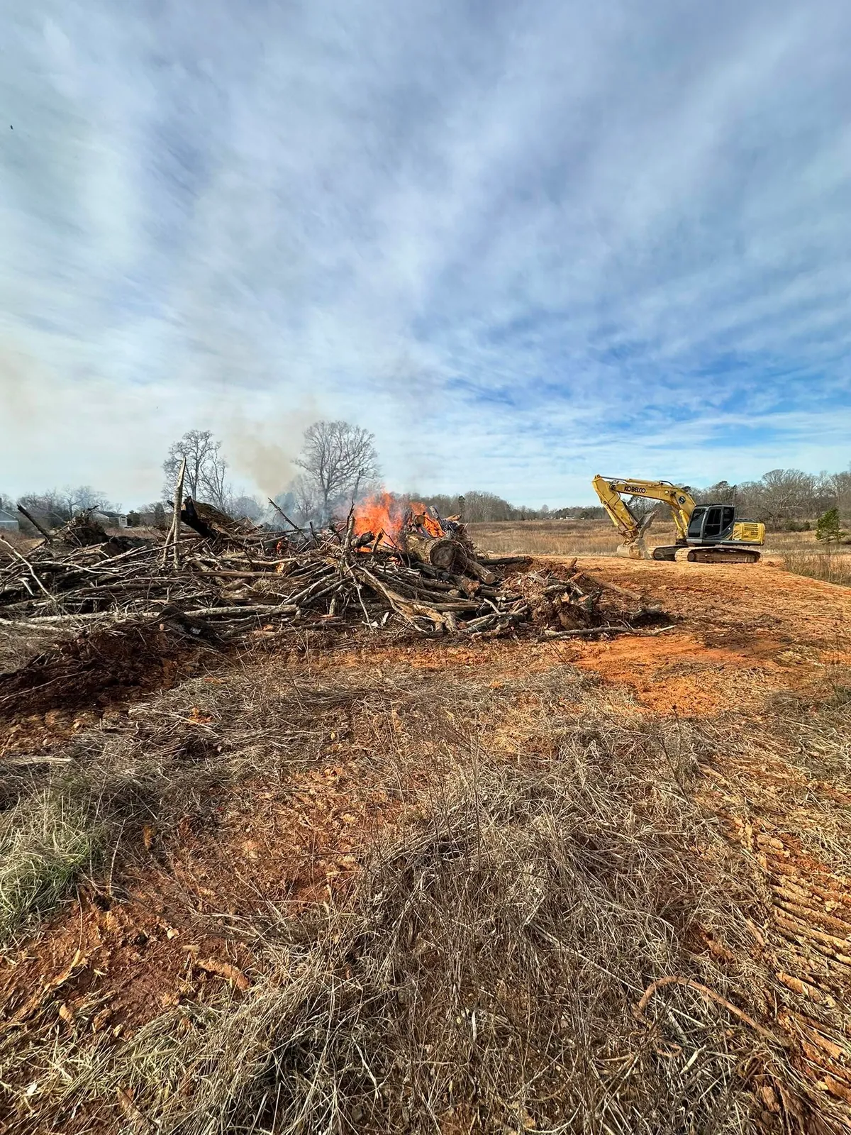 Kobelco excavator beside burning brush pile on cleared red-clay field during land clearing