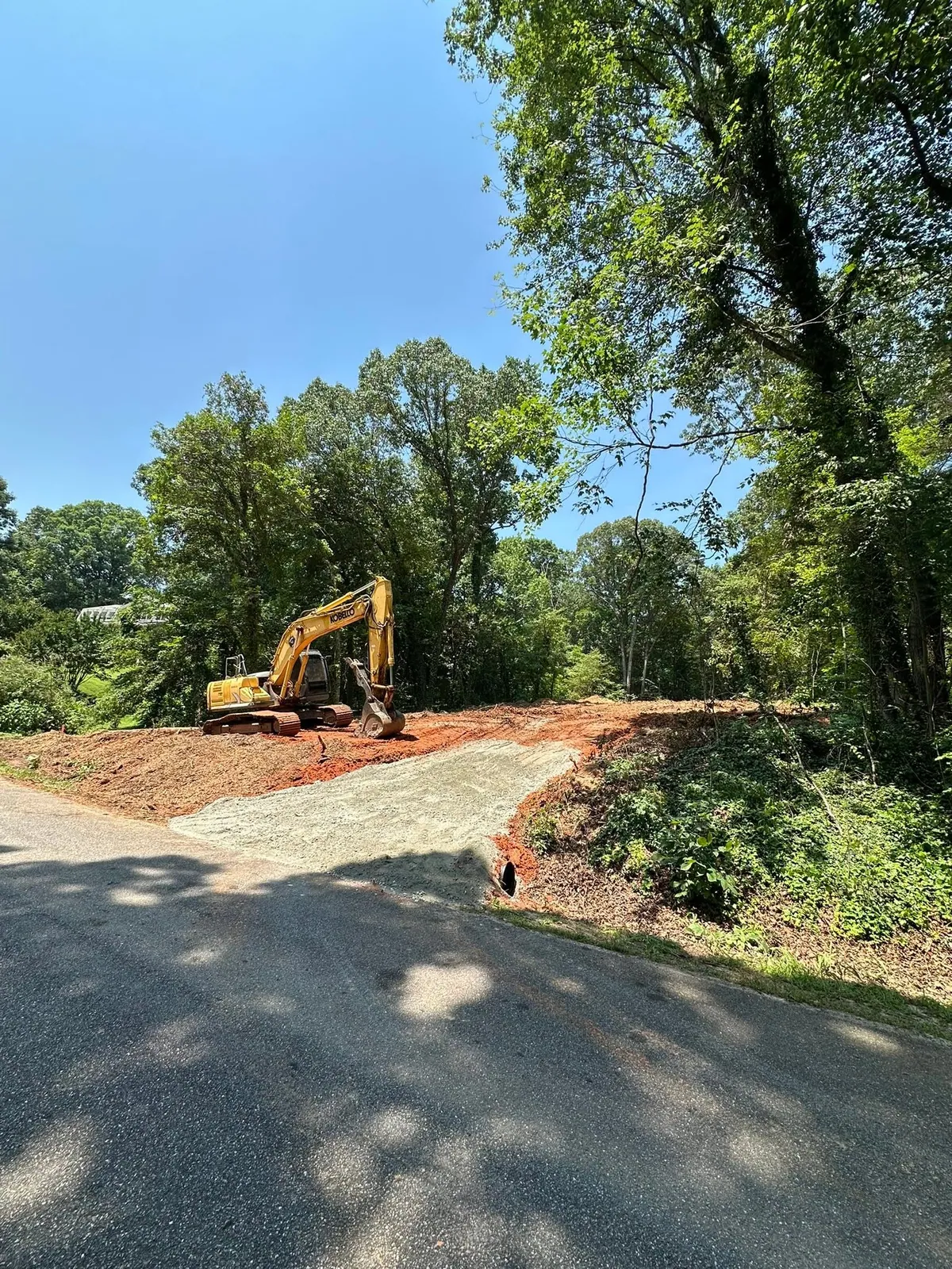 Kobelco excavator beside new gravel driveway entrance with culvert pipe at paved road on cleared lot