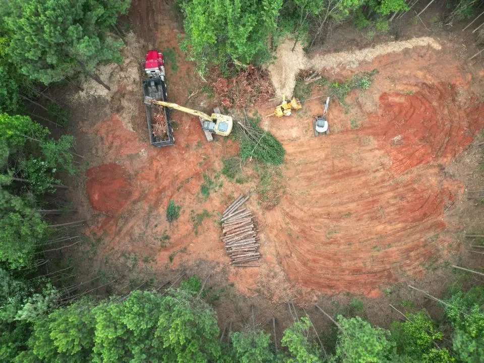 Drone view of excavator loading brush into red dump truck on freshly cleared red-clay wooded lot